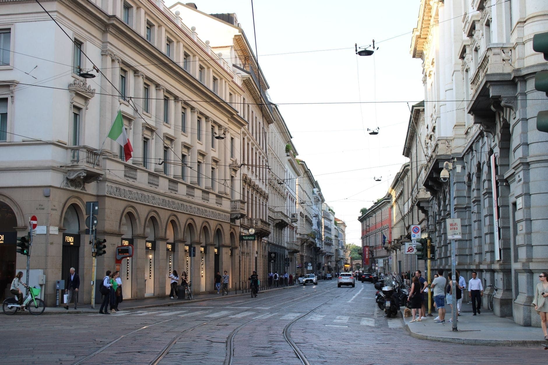 Breite Kopfsteinpflasterstraße in Mailand, gesäumt von eleganten Gebäuden, italienischen Flaggen, Straßenbahnschienen und Fußgängern, die das Stadtzentrum erkunden.