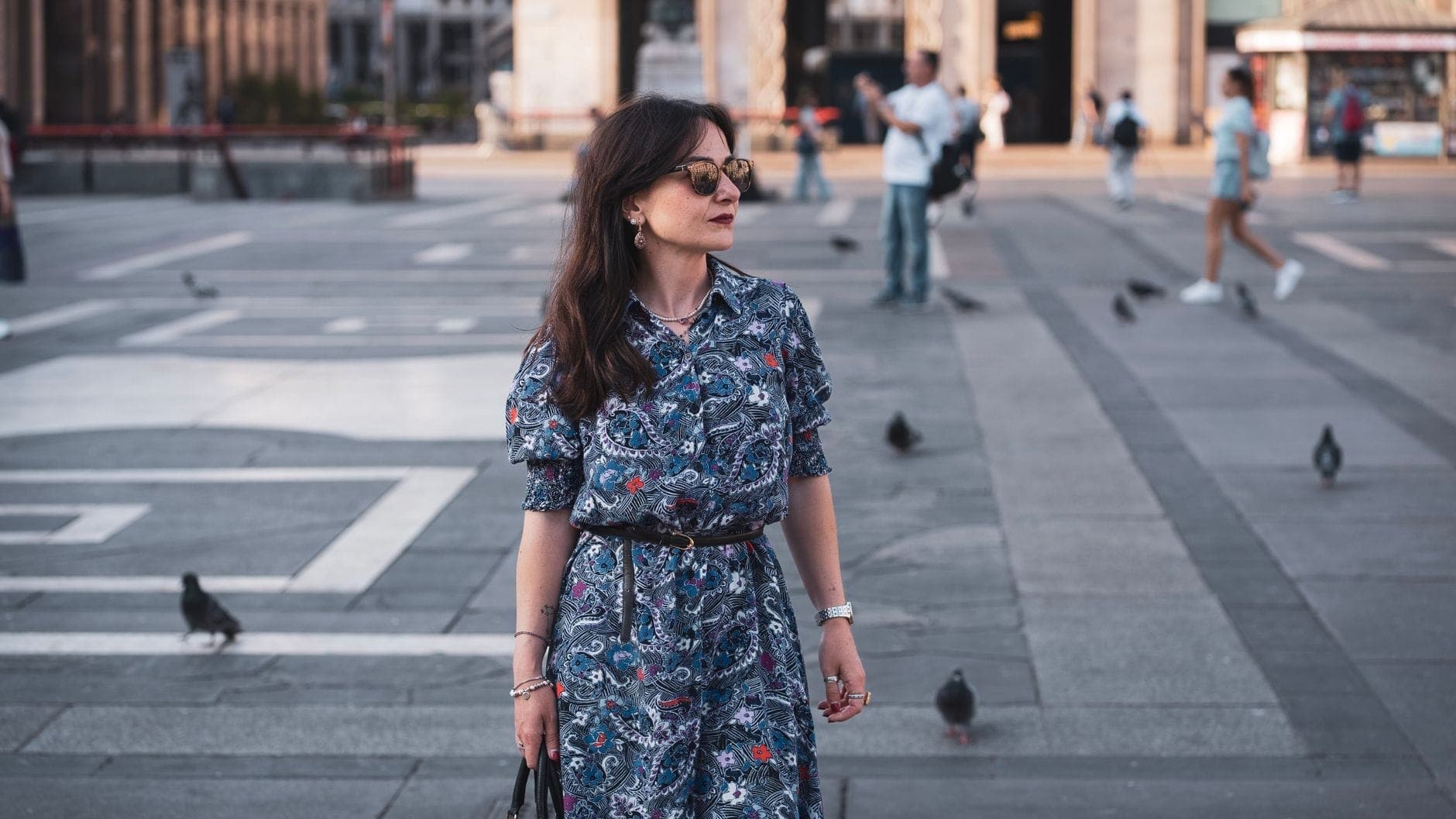 ผู้หญิงสวมชุดลายและแว่นกันแดดเดินผ่าน Piazza del Duomo ในมิลาน โดยมีนกพิราบและสถาปัตยกรรมคลาสสิกเป็นฉากหลัง