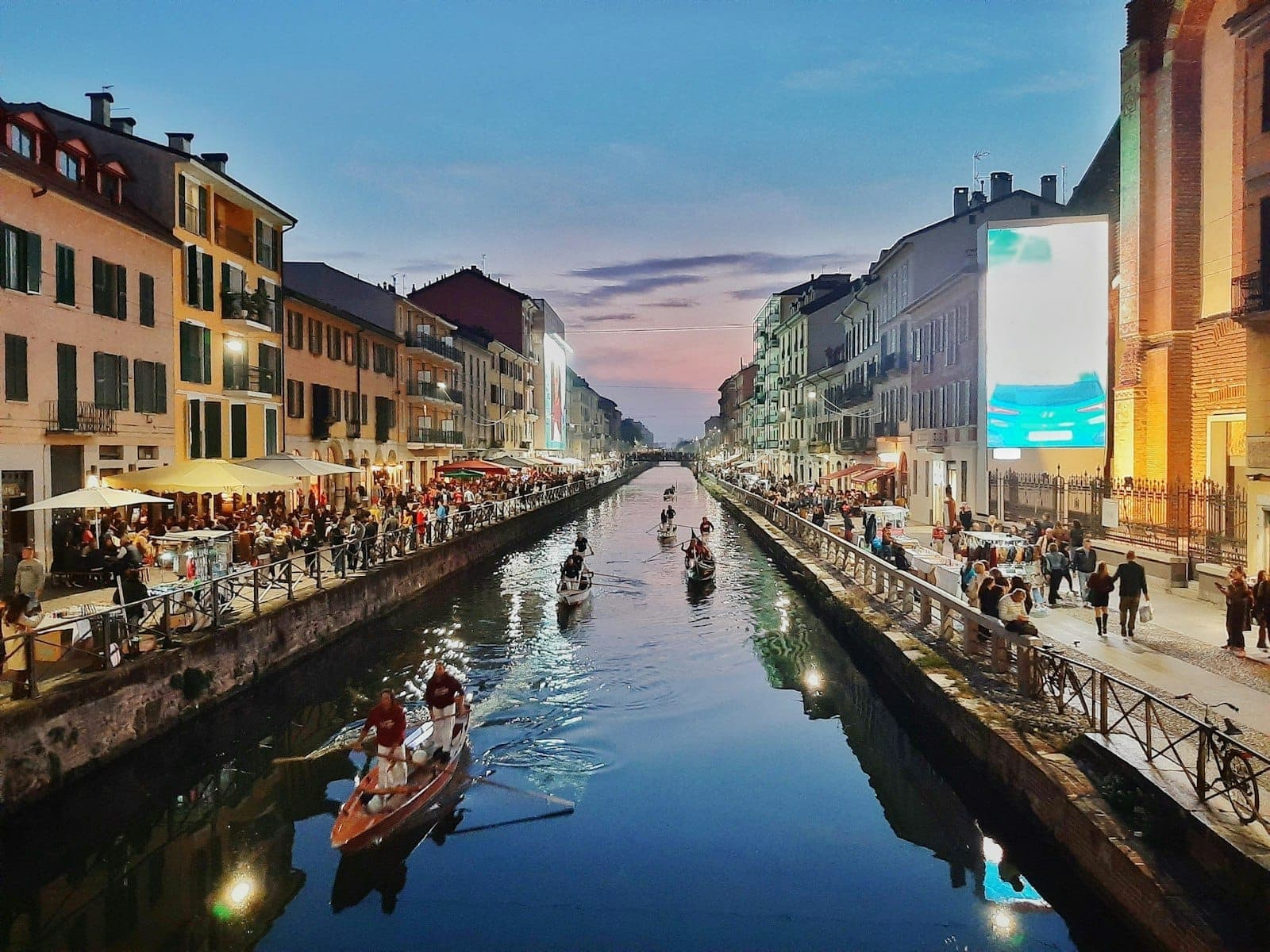 Vista serale del quartiere Navigli di Milano lungo il canale, con ristoranti vivaci, persone che cenano all'aperto e barche sul canale sotto un cielo colorato.