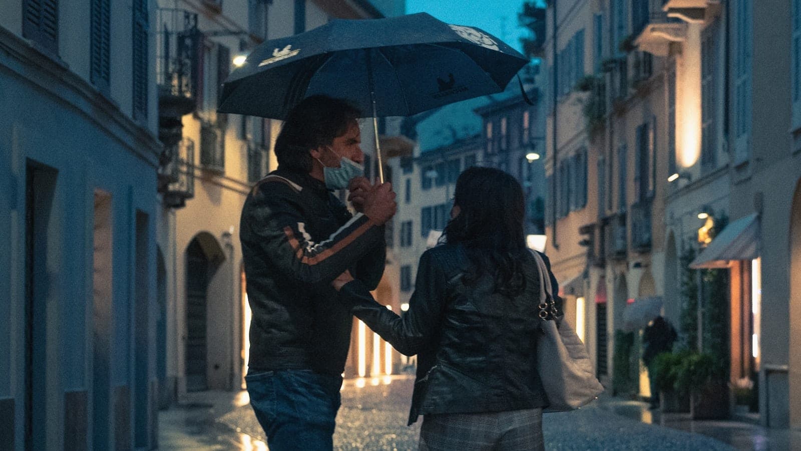 A couple shares an umbrella while walking through a charming cobblestone street in Milan at dusk, softly lit by streetlights.