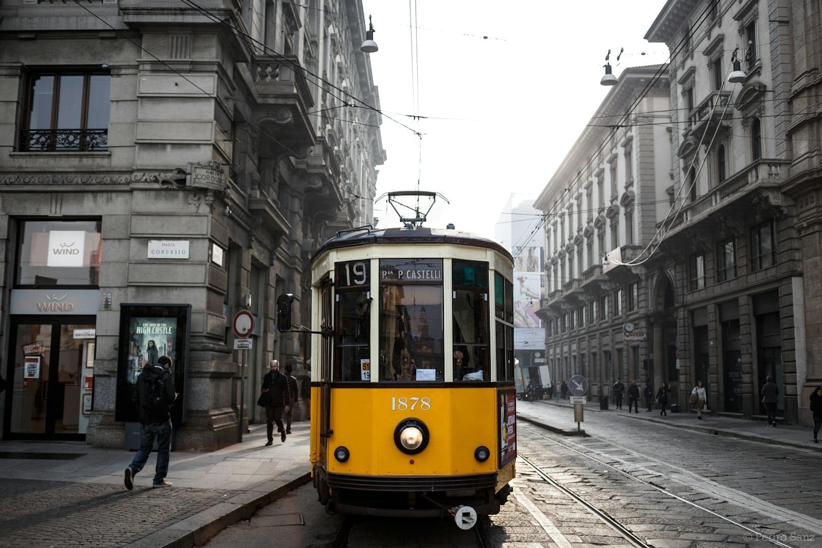 Classic yellow tram number 19 traveling down a narrow street lined with historic stone buildings in central Milan on a misty morning.