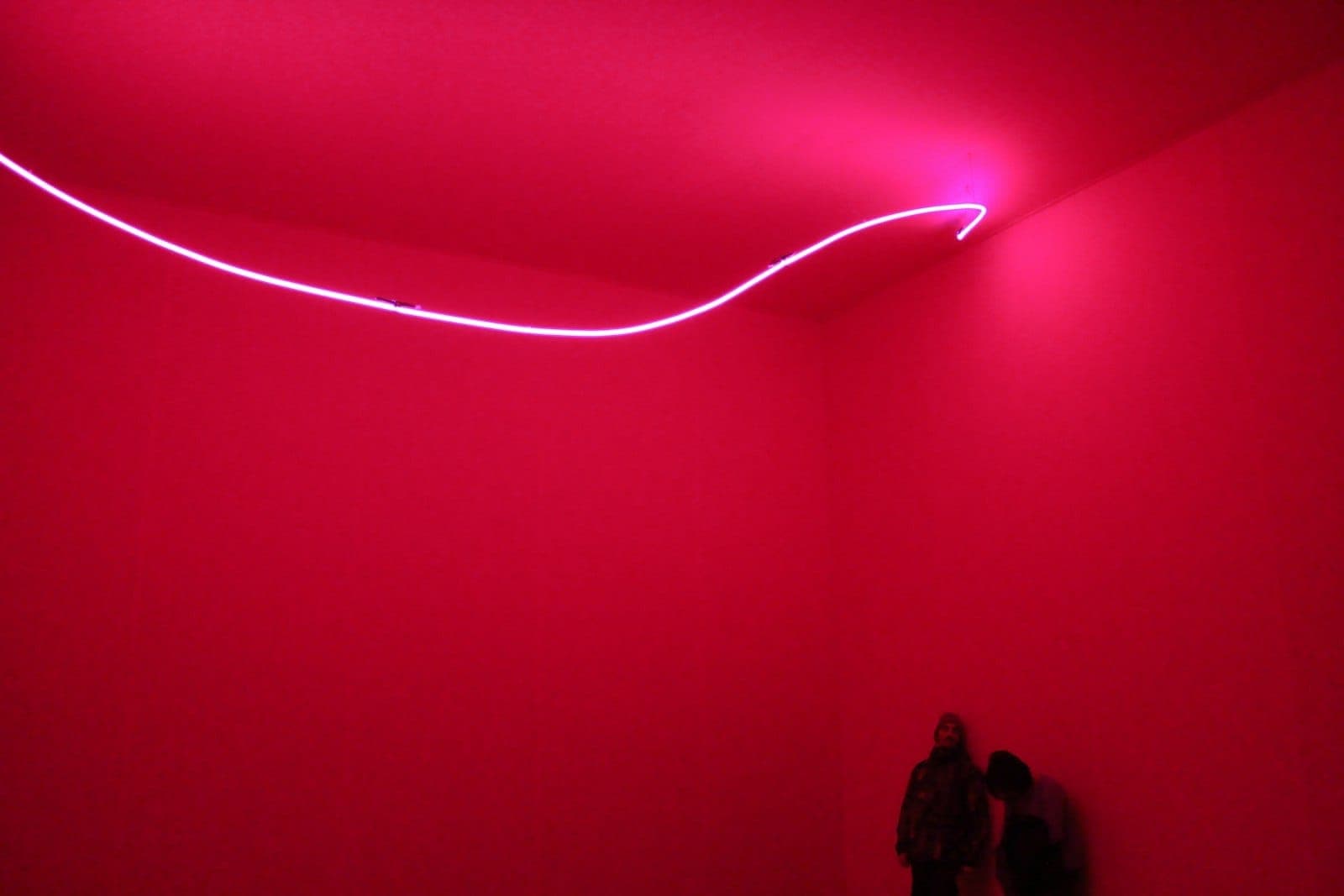 Eine zeitgenössische Kunstinstallation mit einem neonfarbenen rosa Licht in einem minimalistischen, leuchtend roten Raum, zwei Figuren stehen in der Ecke
