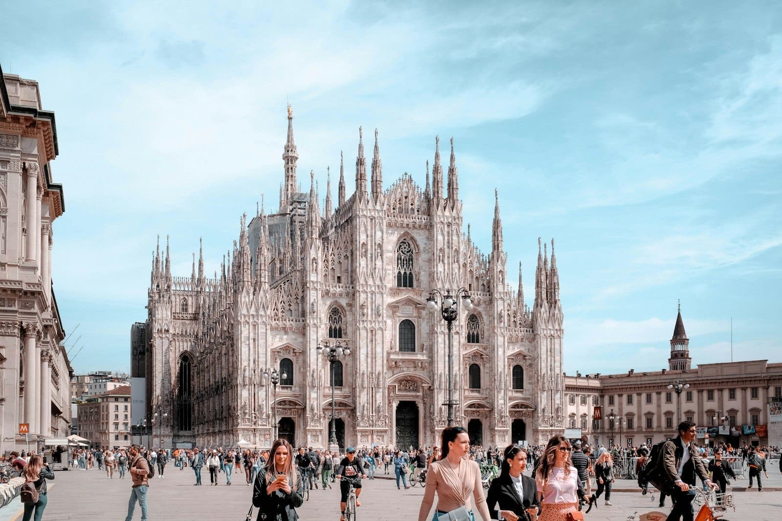 People walking in Piazza del Duomo on a bright spring day with the Milan Cathedral in the background under a blue sky.