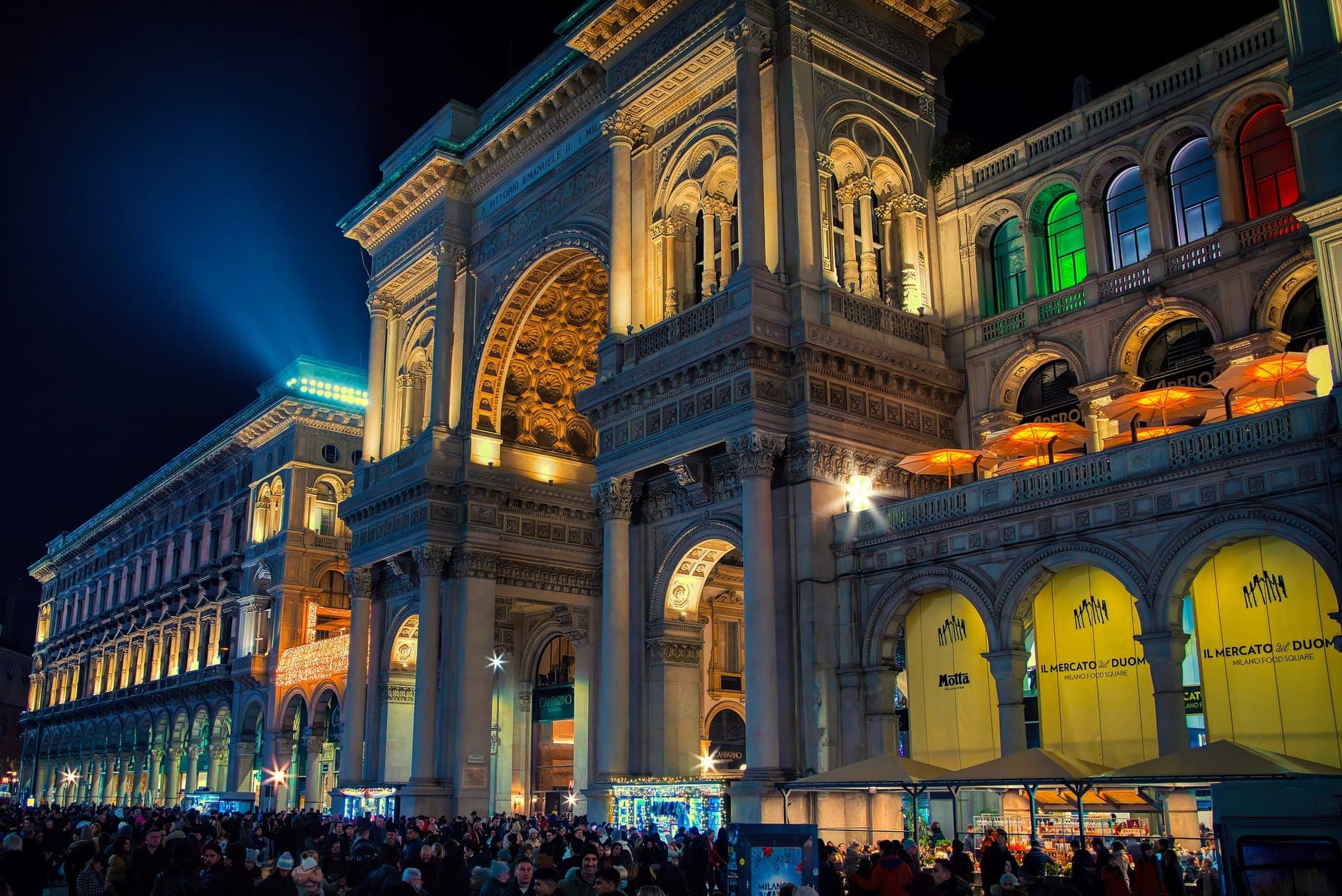 Vibrant night scene at Milan's Galleria Vittorio Emanuele II with crowds enjoying illuminated historic architecture and colorful lights under a dark sky.