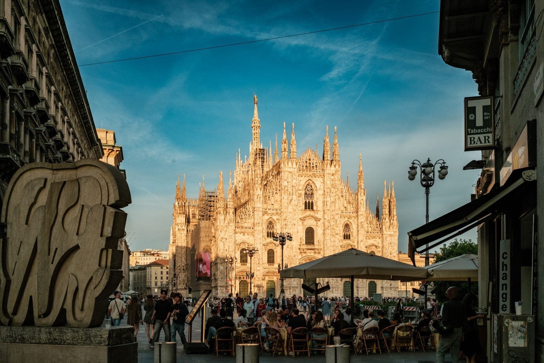 View of Milan Cathedral with outdoor cafes and people in the foreground under a vibrant sunset sky, capturing both local life and iconic architecture.