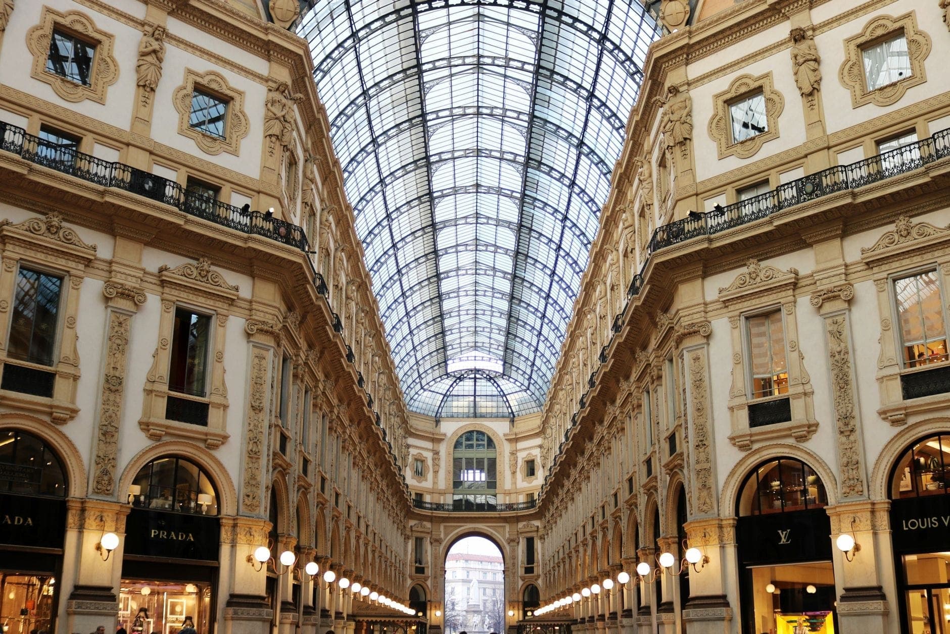 Szeroki widok wnętrza Galleria Vittorio Emanuele II w Mediolanie z ozdobnymi fasadami, szklanym sklepieniem kopułowym i szyldami luksusowych butików po obu stronach.