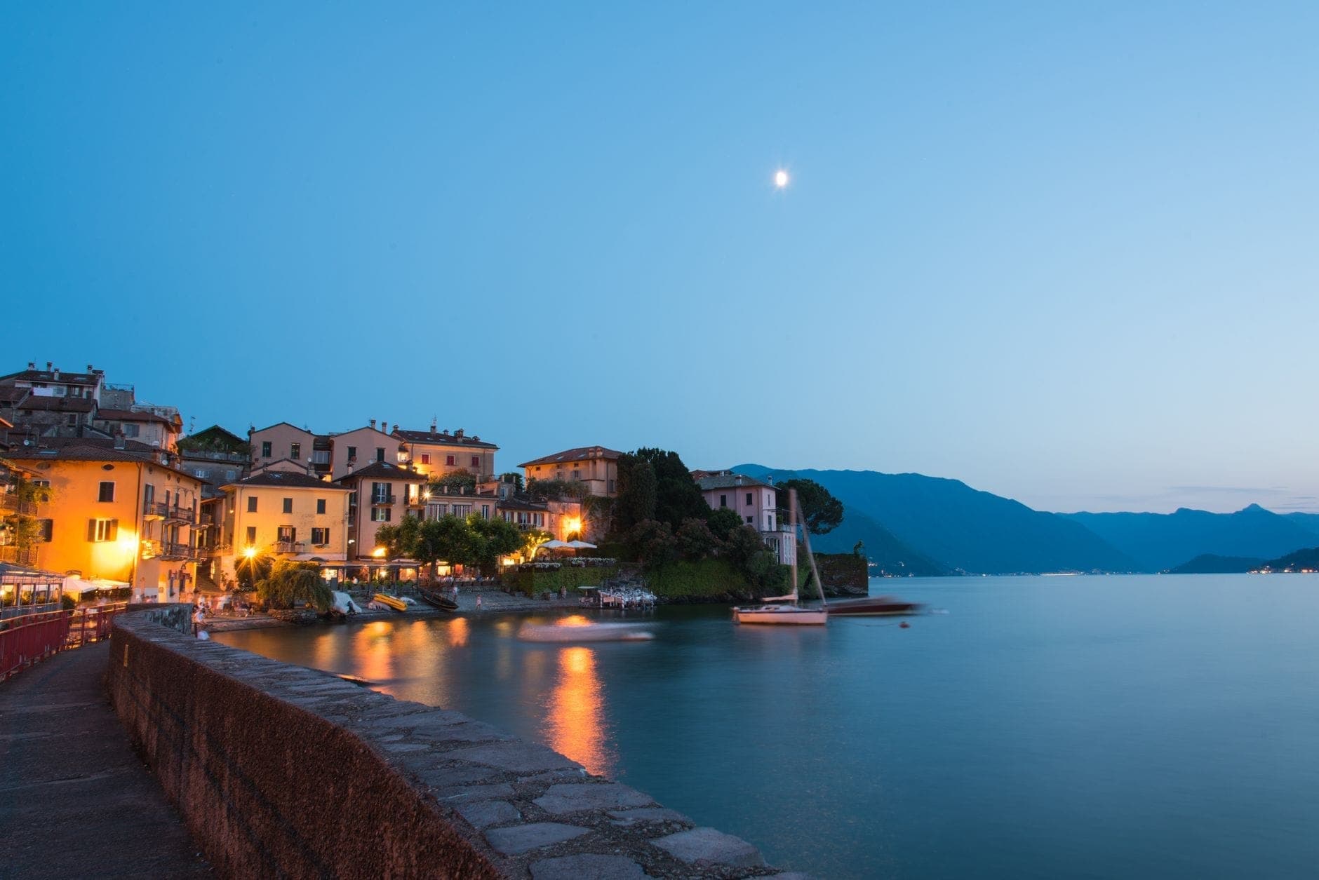 Lake Como waterfront village at dusk with glowing lights reflecting on water, sailboat moored, and mountains in the background under a twilight sky.