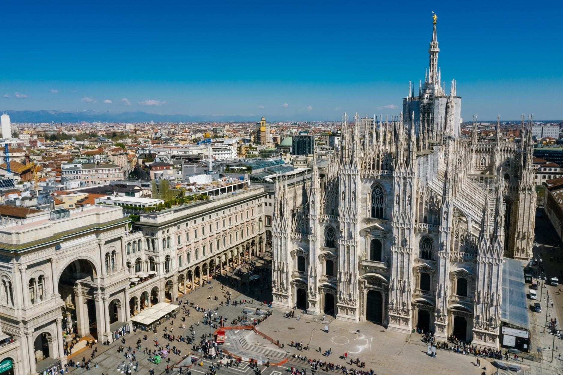 Milano'nun ikonik Duomo katedrali ve çevresindeki şehrin, açık gökyüzü ve aşağıdaki kalabalık piazza ile havadan görünümü.