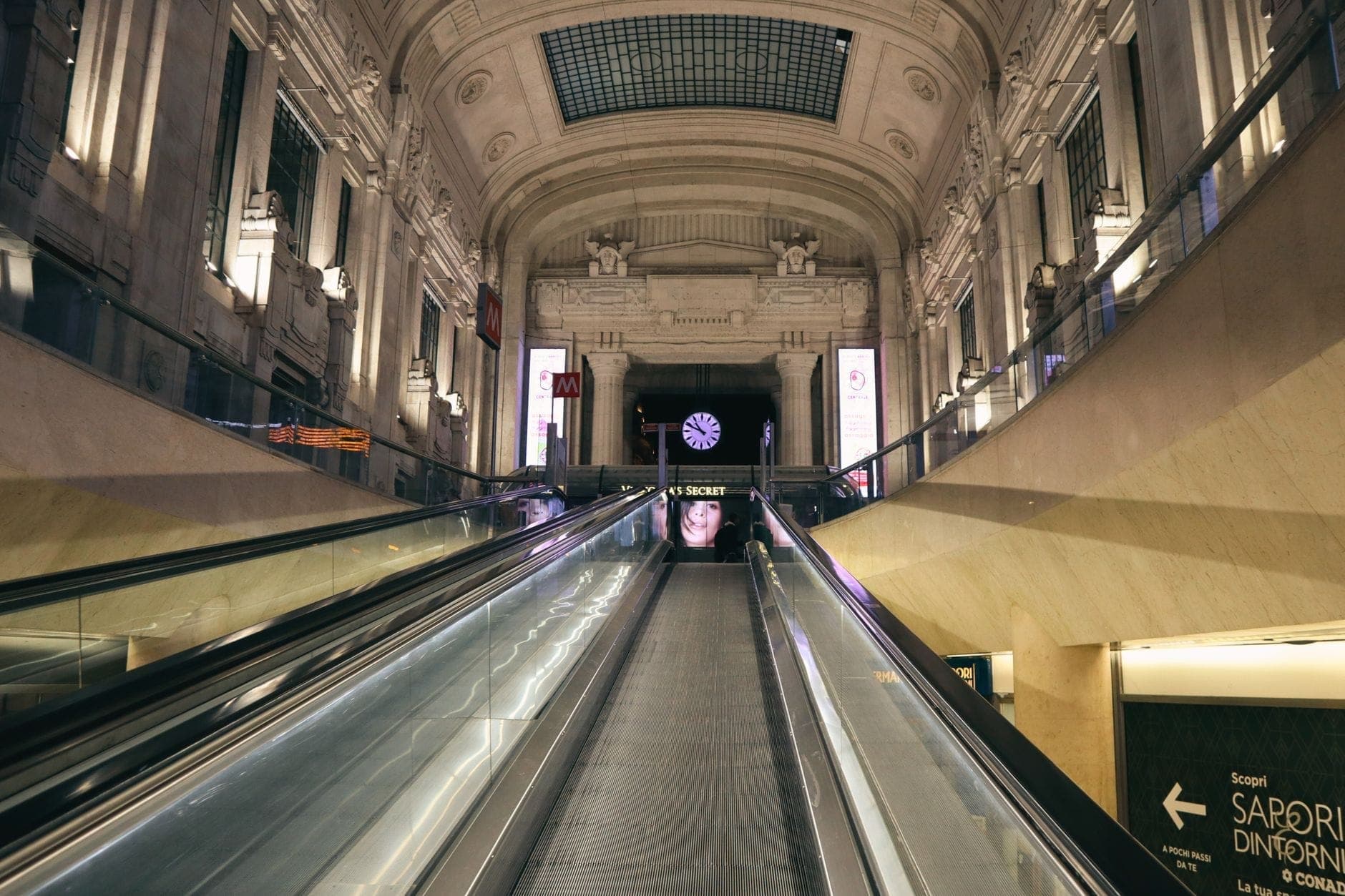 Milano Centrale istasyonunun içindeki bir yürüyen merdivenden yukarı doğru bakış; kemerler, sütunlar ve girişin üzerindeki bir saatiyle görkemli mimariyi sergiliyor.