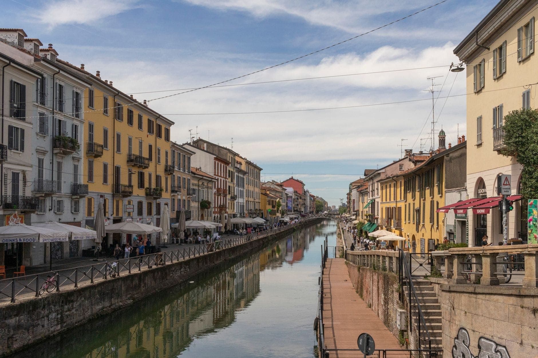 Widok w ciągu dnia na obszar Navigli w Mediolanie z kolorowymi budynkami, kawiarniami na świeżym powietrzu, ożywionymi chodnikami i bezchmurnym niebem ukazującym sezonową aktywność.