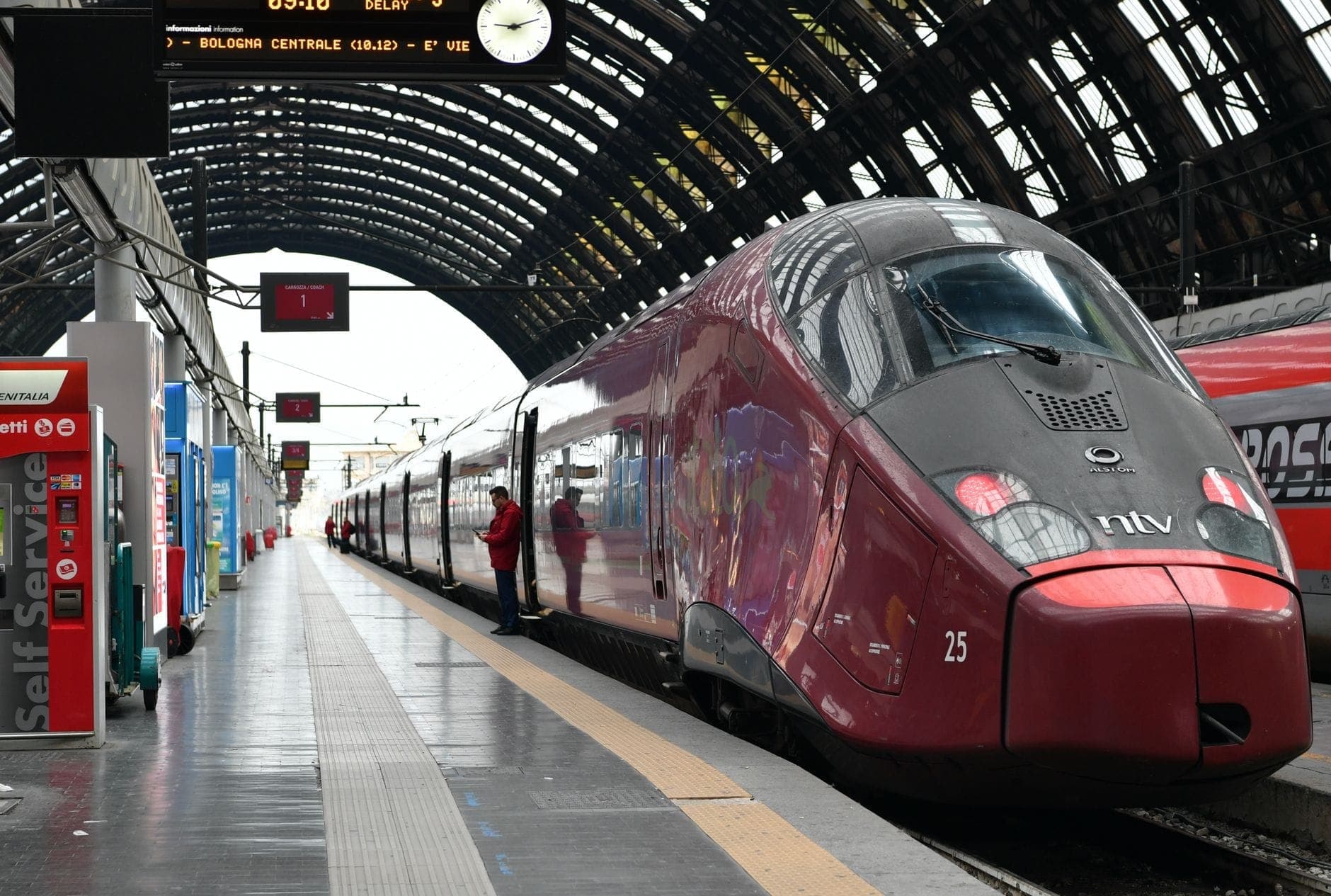 High-speed red Italo train waiting at the platform inside Milan’s grand arched central railway station, with a traveler standing beside the train.