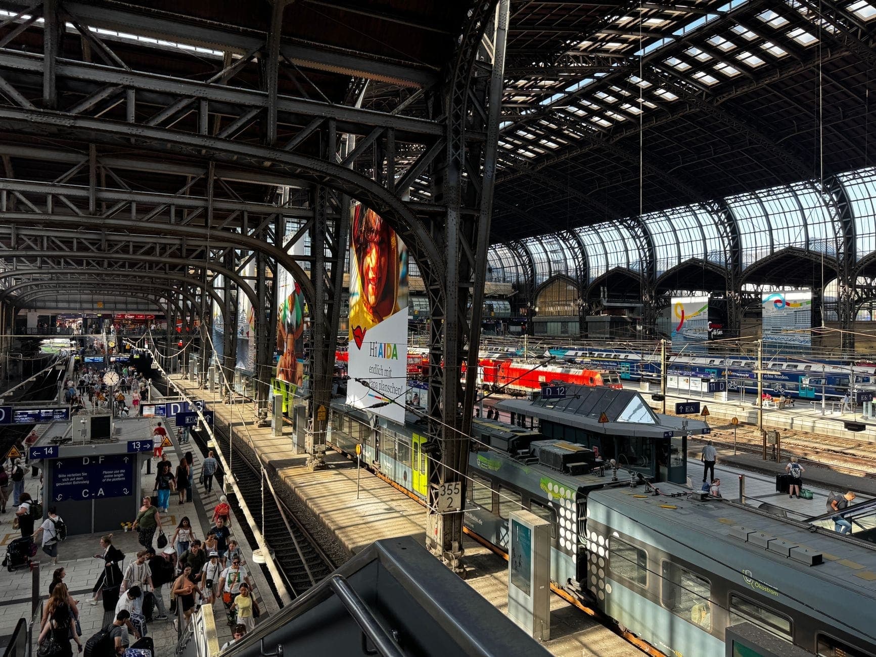 Szeroki widok wnętrza dworca kolejowego Milano Centrale, z pociągami na torach i ludźmi spacerującymi po peronach pod łukowatym szklanym dachem.