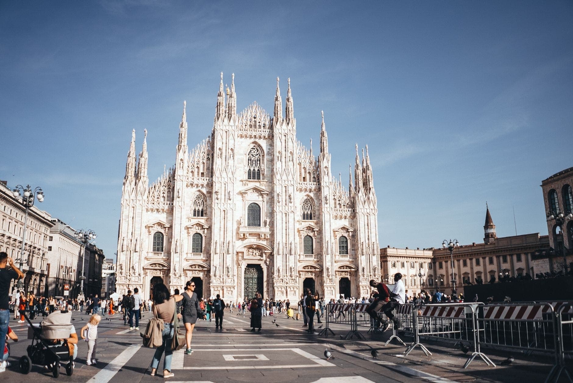 Busy Piazza del Duomo in Milan with families, children in strollers, and tourists walking in front of the cathedral on a sunny day.
