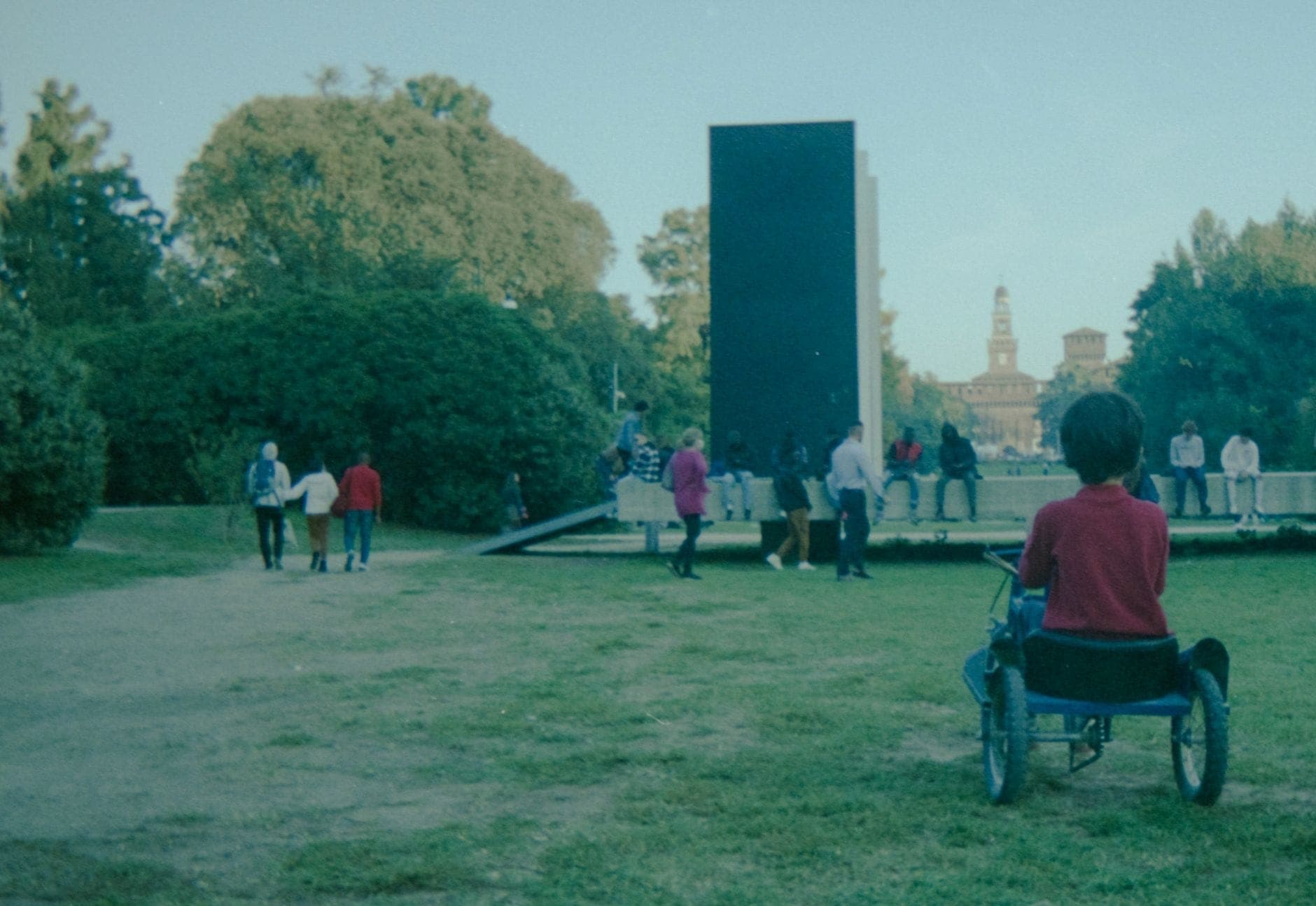 Niños y familias jugando en un parque verde en Milán, con el icónico Castello Sforzesco visible al fondo.