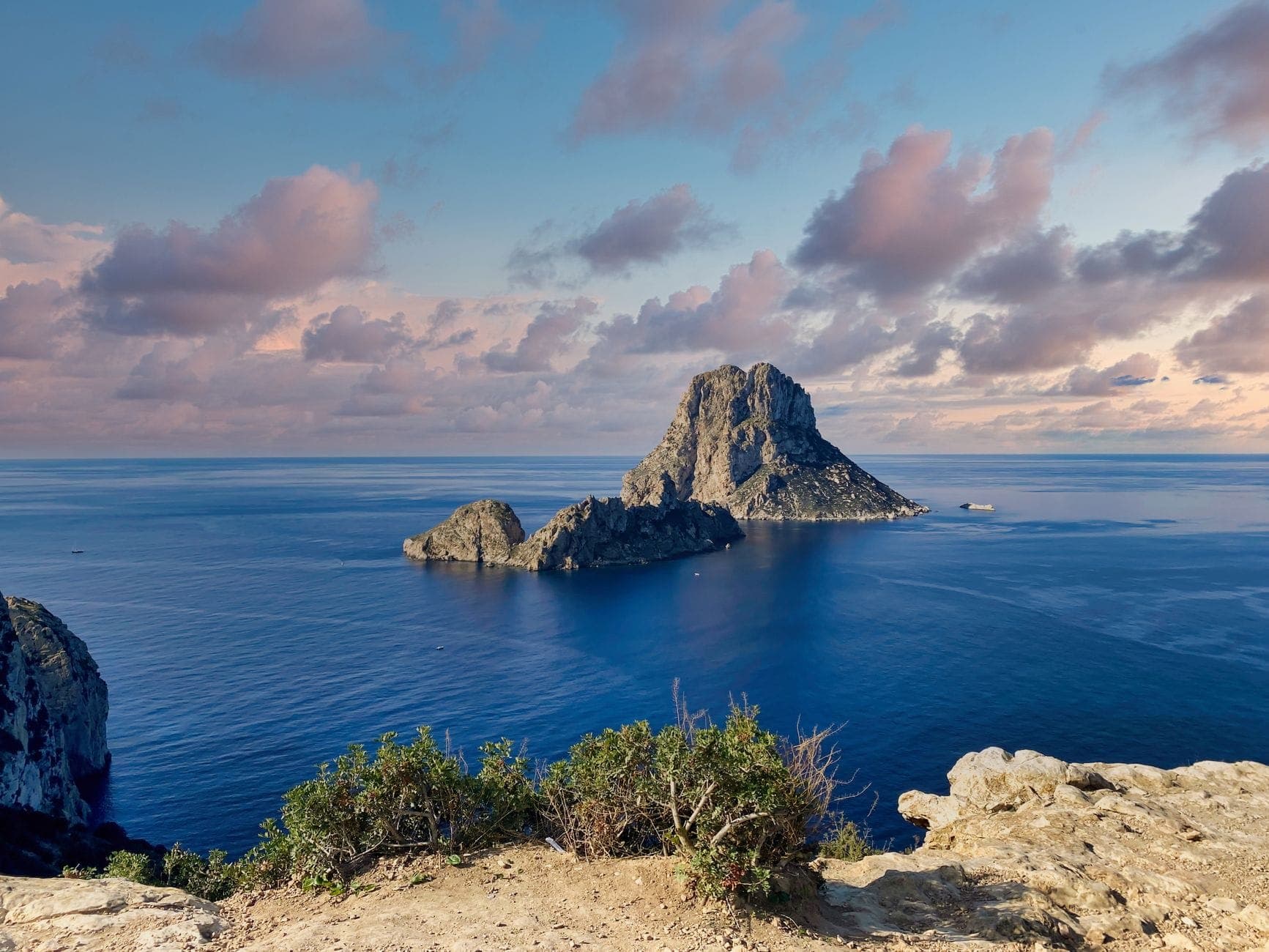 Wide landscape shot of Es Vedrà island off the coast of Ibiza, viewed from Mirador des Vedrà cliffs, with blue sea and dramatic clouds at sunset.