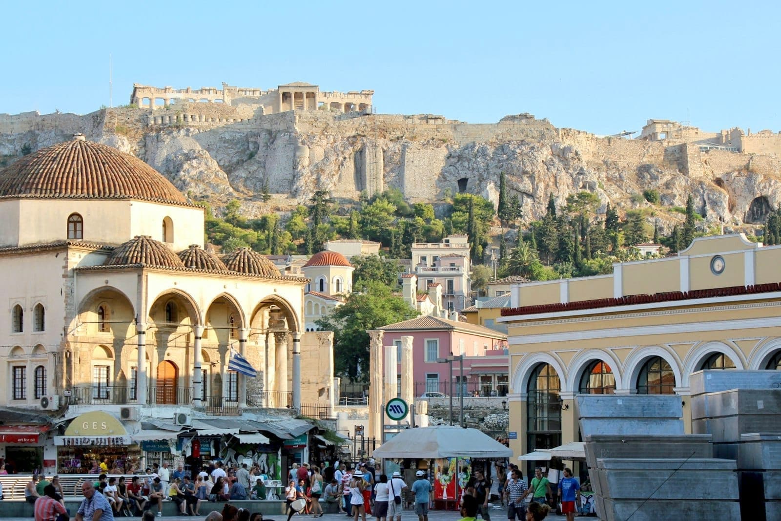Crowds gather in Monastiraki Square with the Tzistarakis Mosque, metro station, and the Acropolis ruins visible in the background on a sunny day.