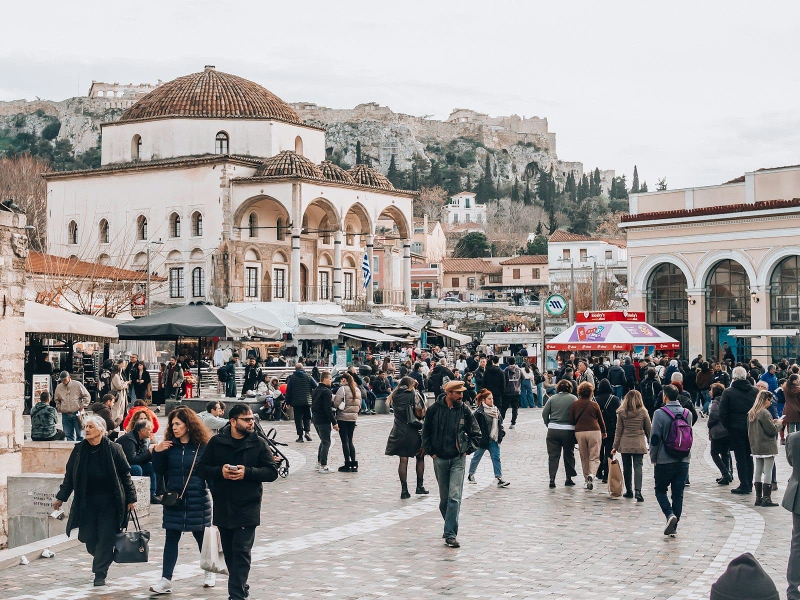Crowds explore Monastiraki Flea Market in Athens, surrounded by historic buildings and stalls, with the Acropolis visible in the background.