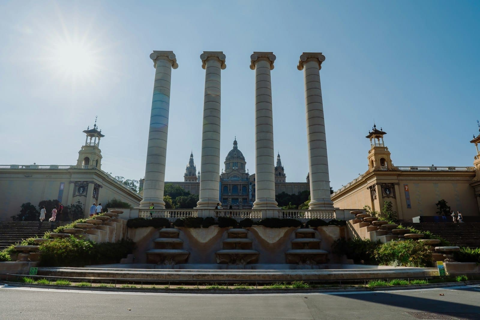 Tanawin ng apat na haligi kasama ang Magic Fountain at ang Pambansang Museo ng Sining ng Catalonia sa likod, sa ilalim ng maliwanag na asul na langit sa Montjuïc, Barcelona.