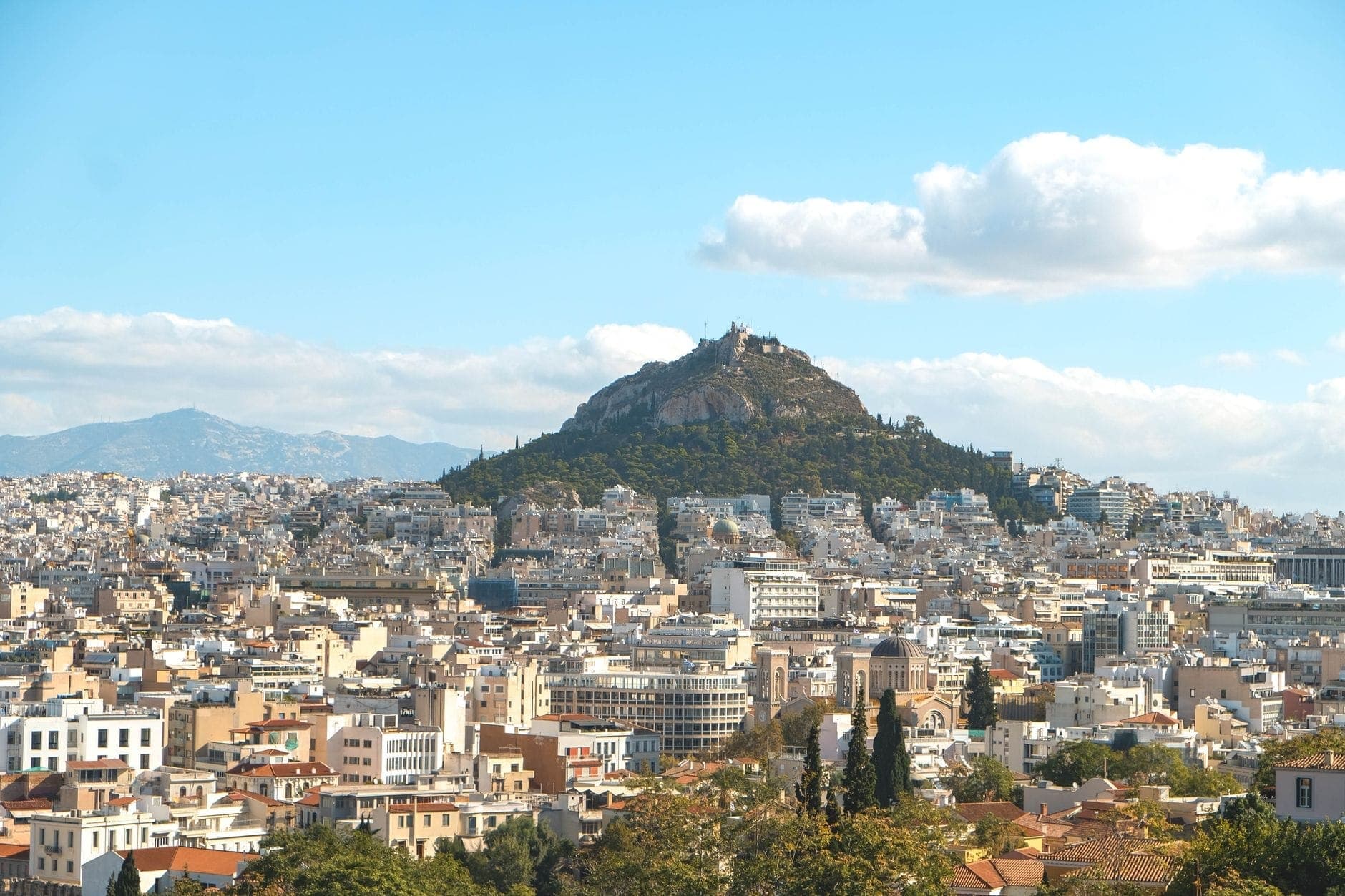 Mount Lycabettus rises above the cityscape of Athens under a bright blue sky with soft clouds, offering a panoramic and inviting view for travelers.
