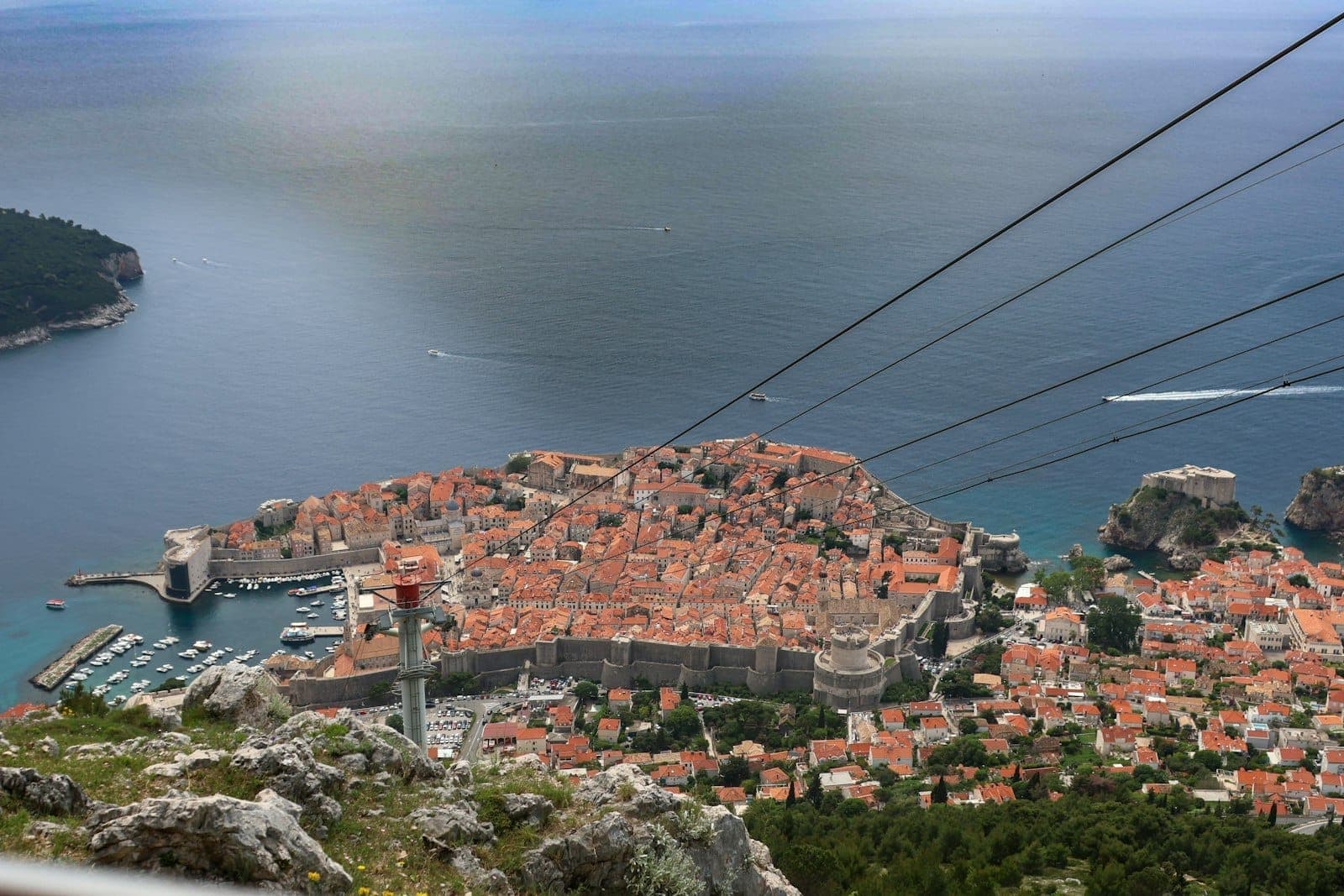Panoramikong tanawin mula sa Mount Srđ na nagpapakita ng Lumang Lungsod ng Dubrovnik, Dagat Adriatiko, Isla ng Lokrum, at mga kawad ng cable car na bumababa patungo sa lungsod sa ibaba.