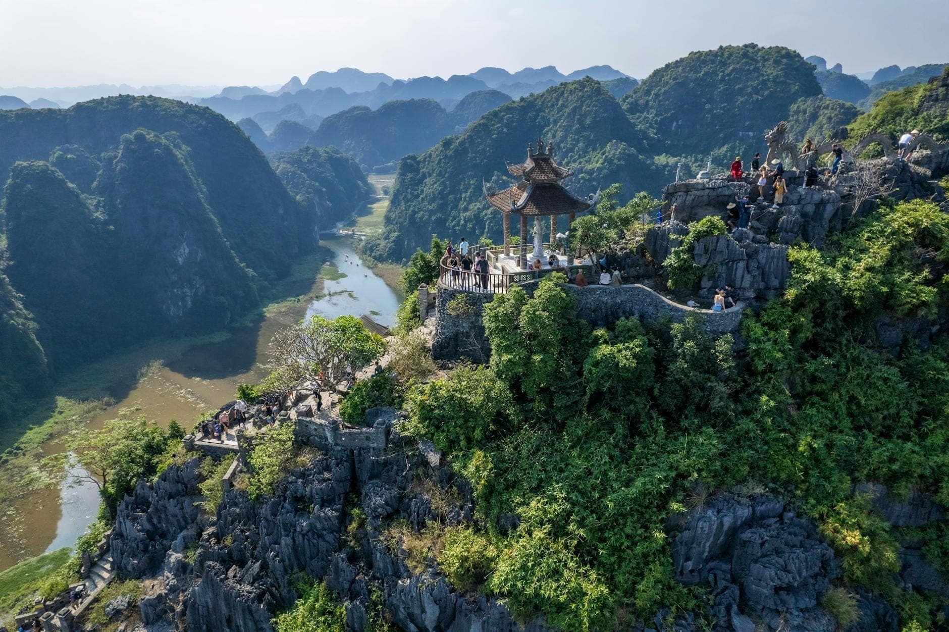 Toàn cảnh đỉnh Hang Mua nhìn từ trên cao với du khách tại vọng lâu trên đỉnh, phía dưới là núi đá vôi trùng điệp, dòng sông uốn lượn và những cánh đồng lúa xanh mướt ở Ninh Bình, Việt Nam.