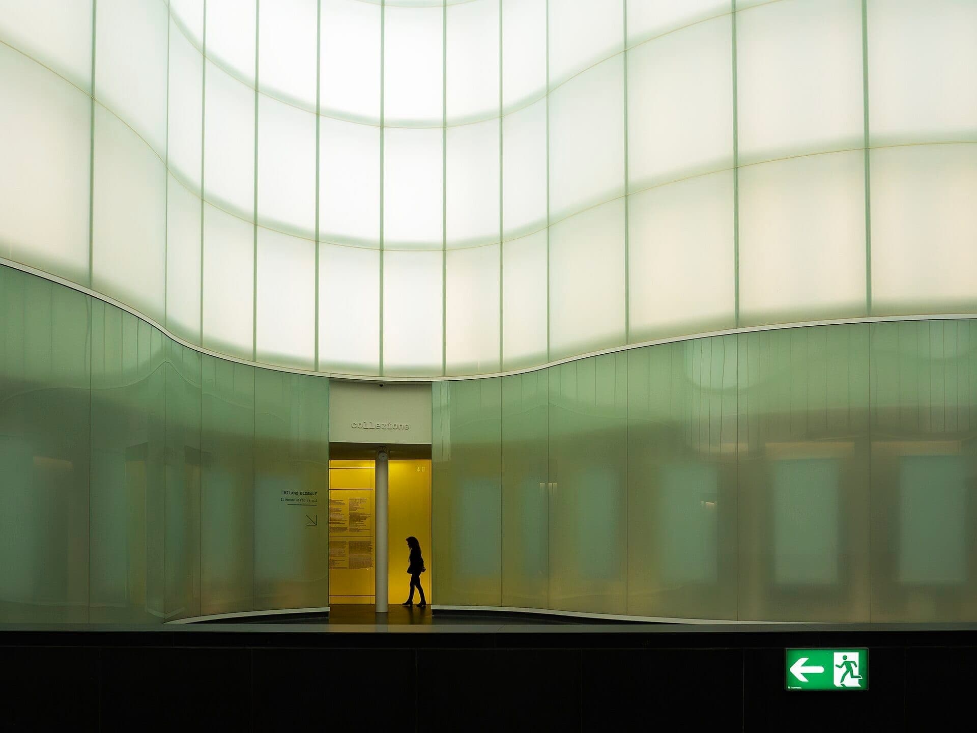 Interior view of MUDEC – Museo delle Culture in Milan, featuring a lone visitor, soft curved glass walls, and glowing modern lighting.