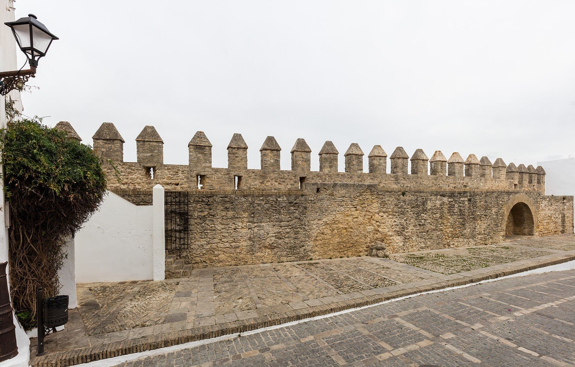 Những tường thành bằng đá của Murallas de Cádiz với lối vào hình vòm lịch sử, lối đi lát đá cuội và ánh sáng dịu nhẹ dưới bầu trời흐리 흐리.