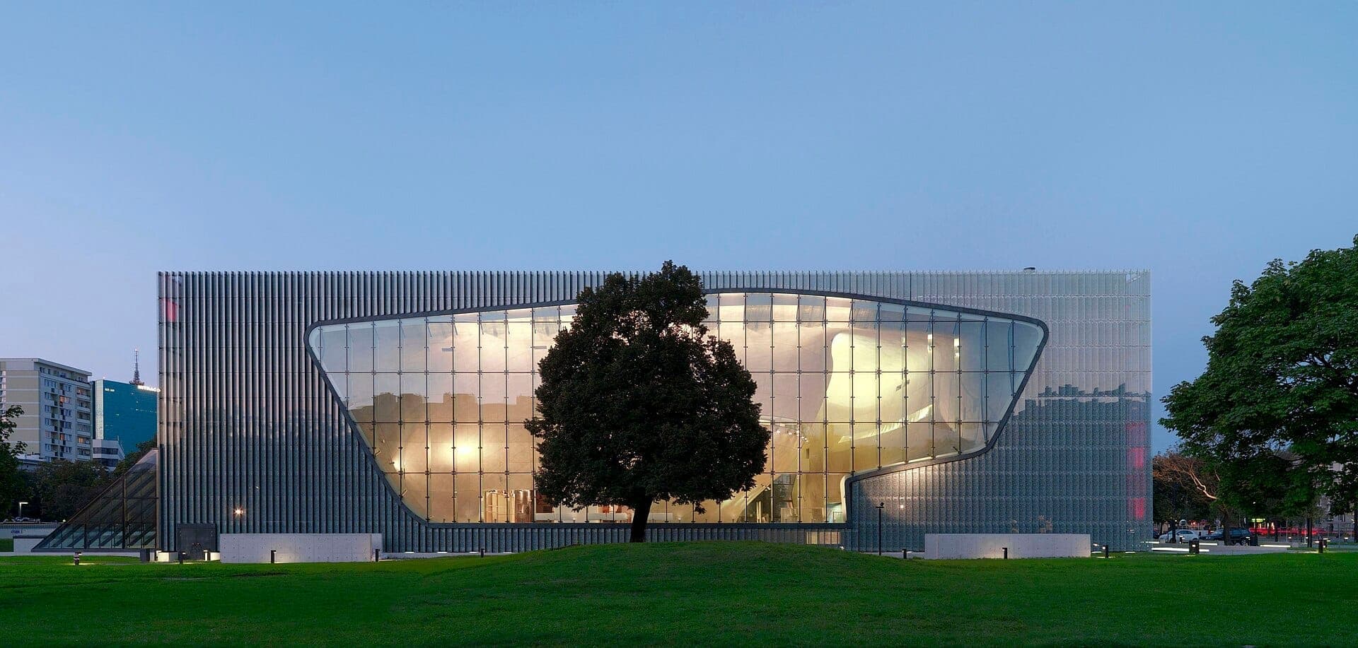 Moderne Glasfassade des POLIN Museums in Muranów, Warschau, in der Abenddämmerung mit beleuchtetem Inneren und grünem Rasen im Vordergrund.