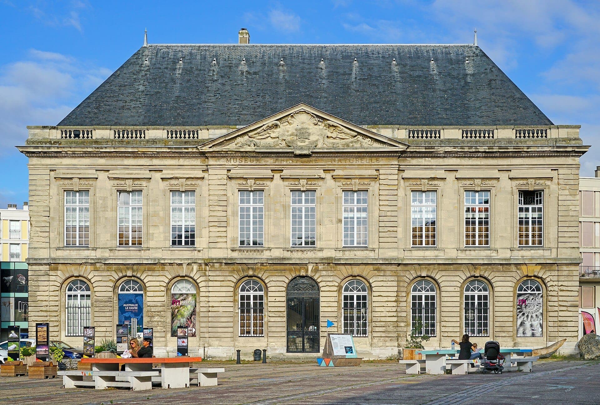 Muséum d'Histoire Naturelle du Havre nổi bật với mặt tiền đá uy nghi, những ô cửa sổ lớn và tam giác sơn hoa, nằm dưới bầu trời xanh.
