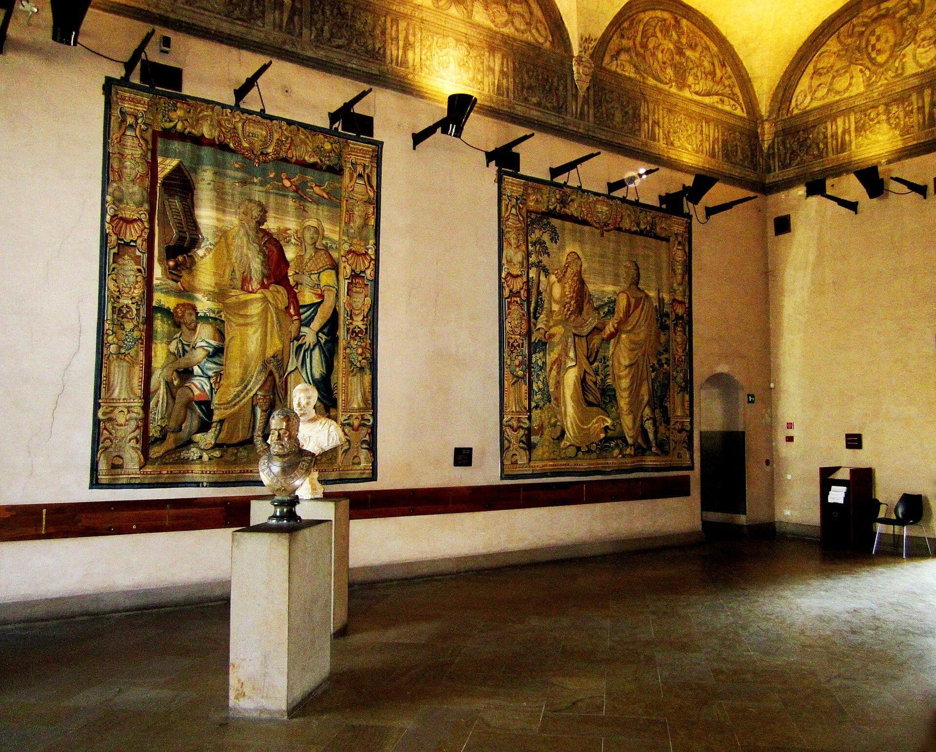 Interior gallery of Musei del Castello Sforzesco with two ornate Renaissance tapestries, marble busts, arched ceiling, and warm lighting.