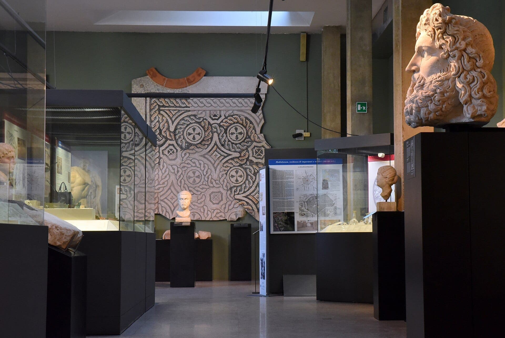 Exhibit hall at Museo Civico Archeologico di Milano showcasing Roman busts, informational panels, and a large decorative mosaic on the wall.