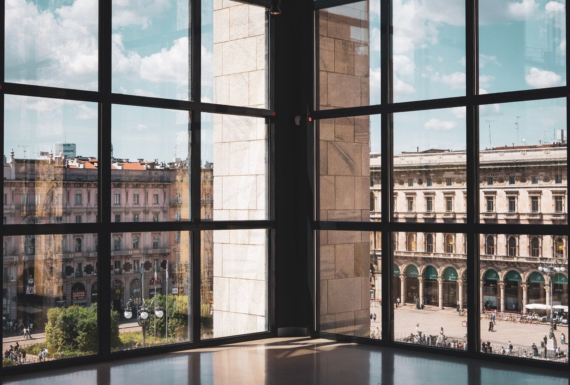 View of Piazza del Duomo and historic buildings through large glass windows at Museo del Novecento in Milan, on a sunny day.