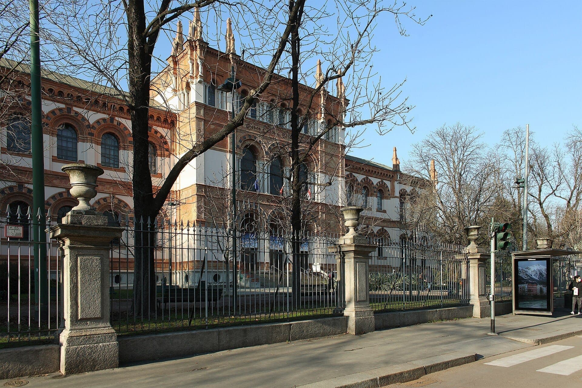 The neo-Gothic facade of Museo di Storia Naturale di Milano with arched windows, iron fence, bare trees, and clear blue sky.