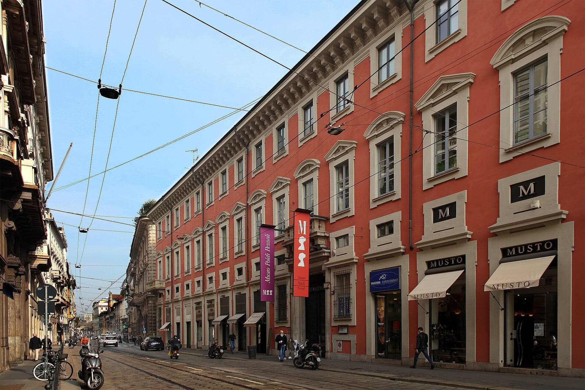 The exterior of Museo Poldi Pezzoli in Milan, featuring a grand coral-colored building along Via Manzoni with people walking by.