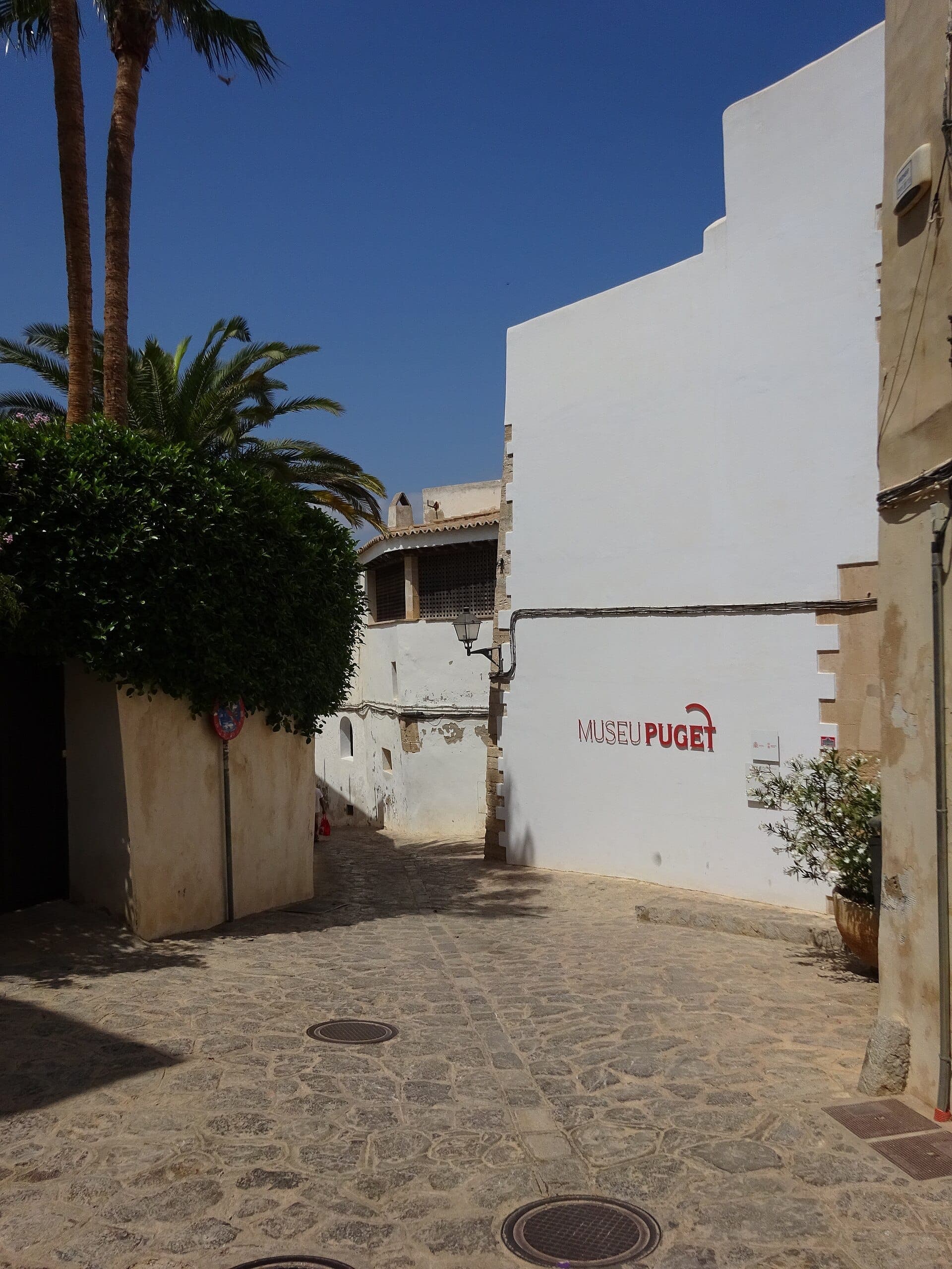 Narrow stone street leading to the whitewashed exterior of Museu Puget in Ibiza, with palm trees and bright blue sky overhead.