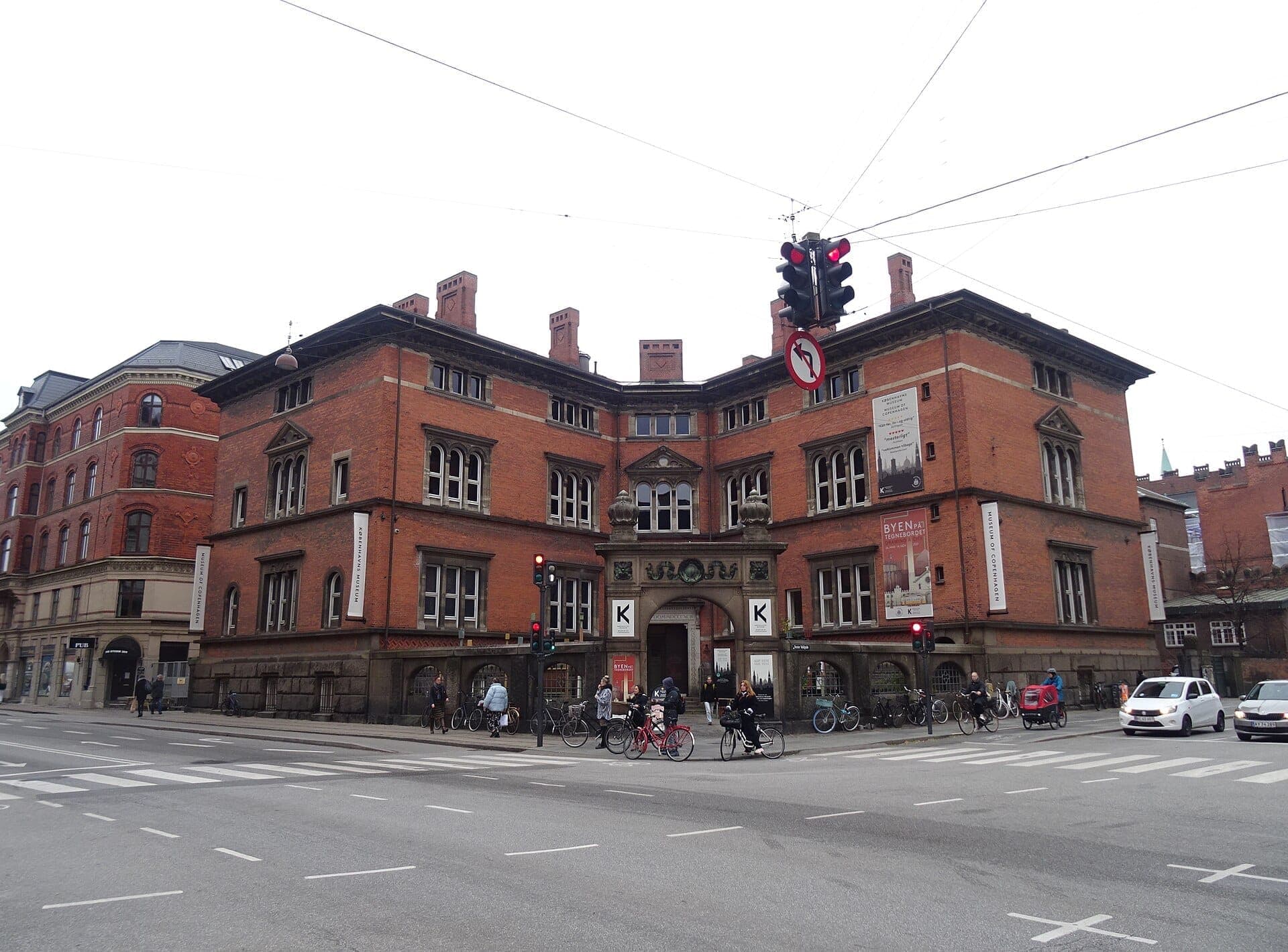 Das rote Backsteingebäude des Museum of Copenhagen an einem bewölkten Tag, mit Radfahrern, Fußgängern und Autos an der belebten Kreuzung davor.