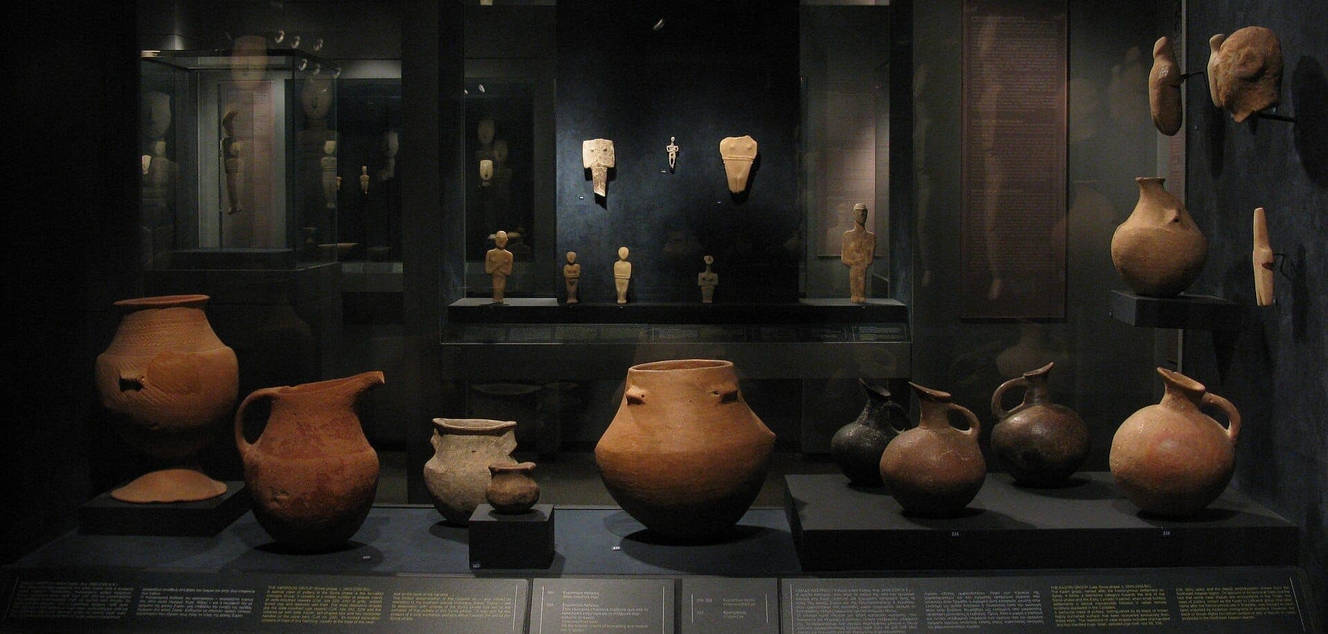 Exhibit at the Museum of Cycladic Art featuring ancient terracotta pots and Cycladic figurines displayed in dimly lit glass cases.