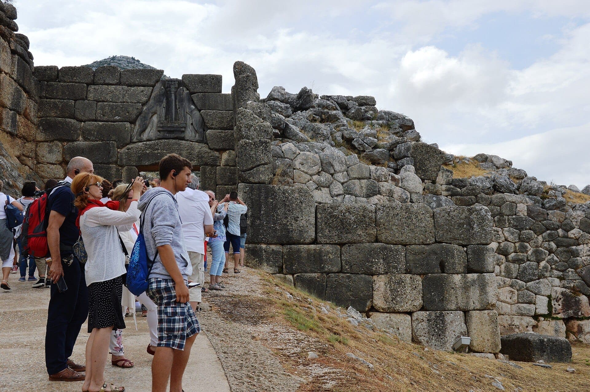 Visitors stand near the ancient Lion Gate, surrounded by massive Cyclopean stone walls at the archaeological site of Mycenae under a cloudy sky.