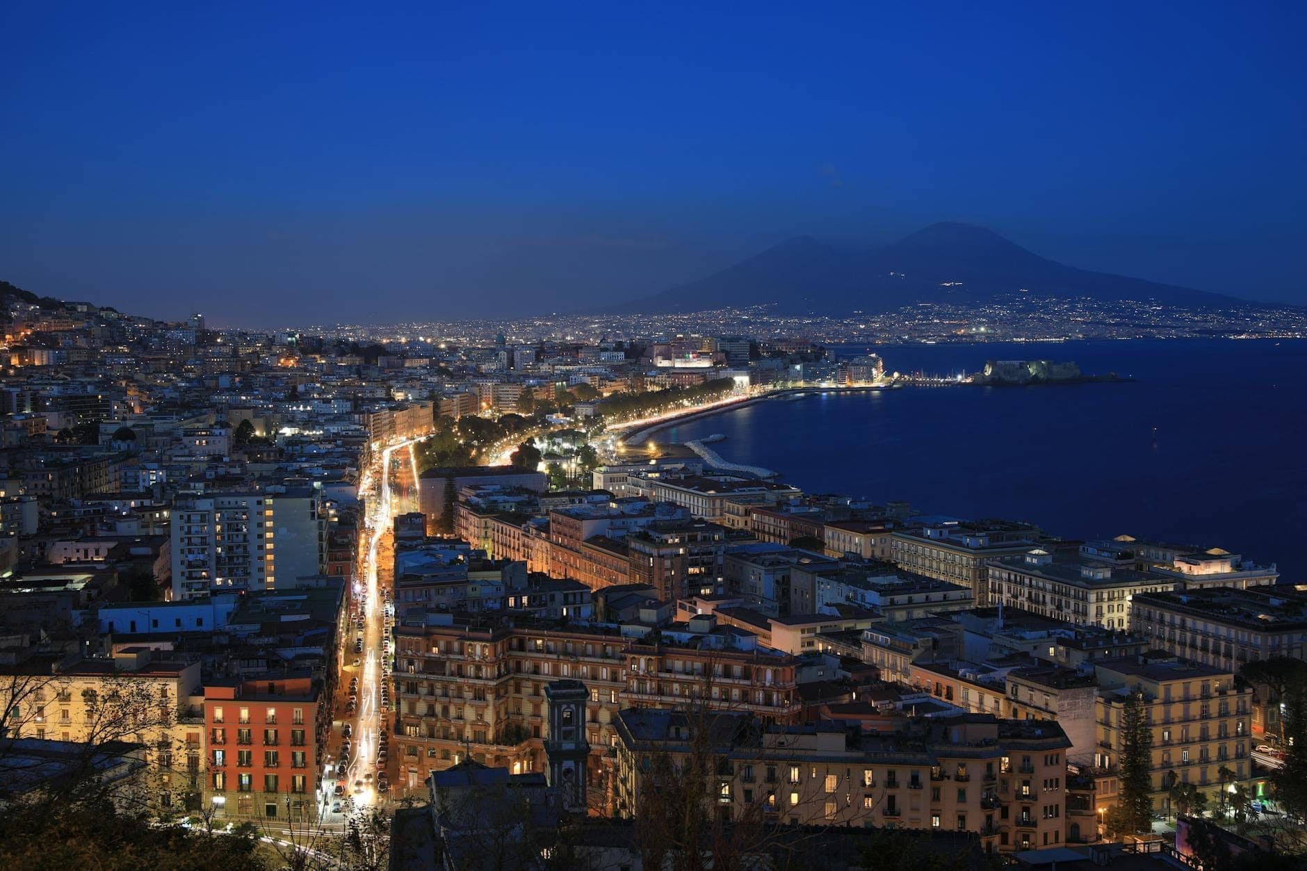Toàn cảnh bờ biển Naples về đêm với ánh đèn thành phố, những con phố nhộn nhịp và núi Vesuvius sừng sững ở phía sau dưới bầu trời xanh thẫm.