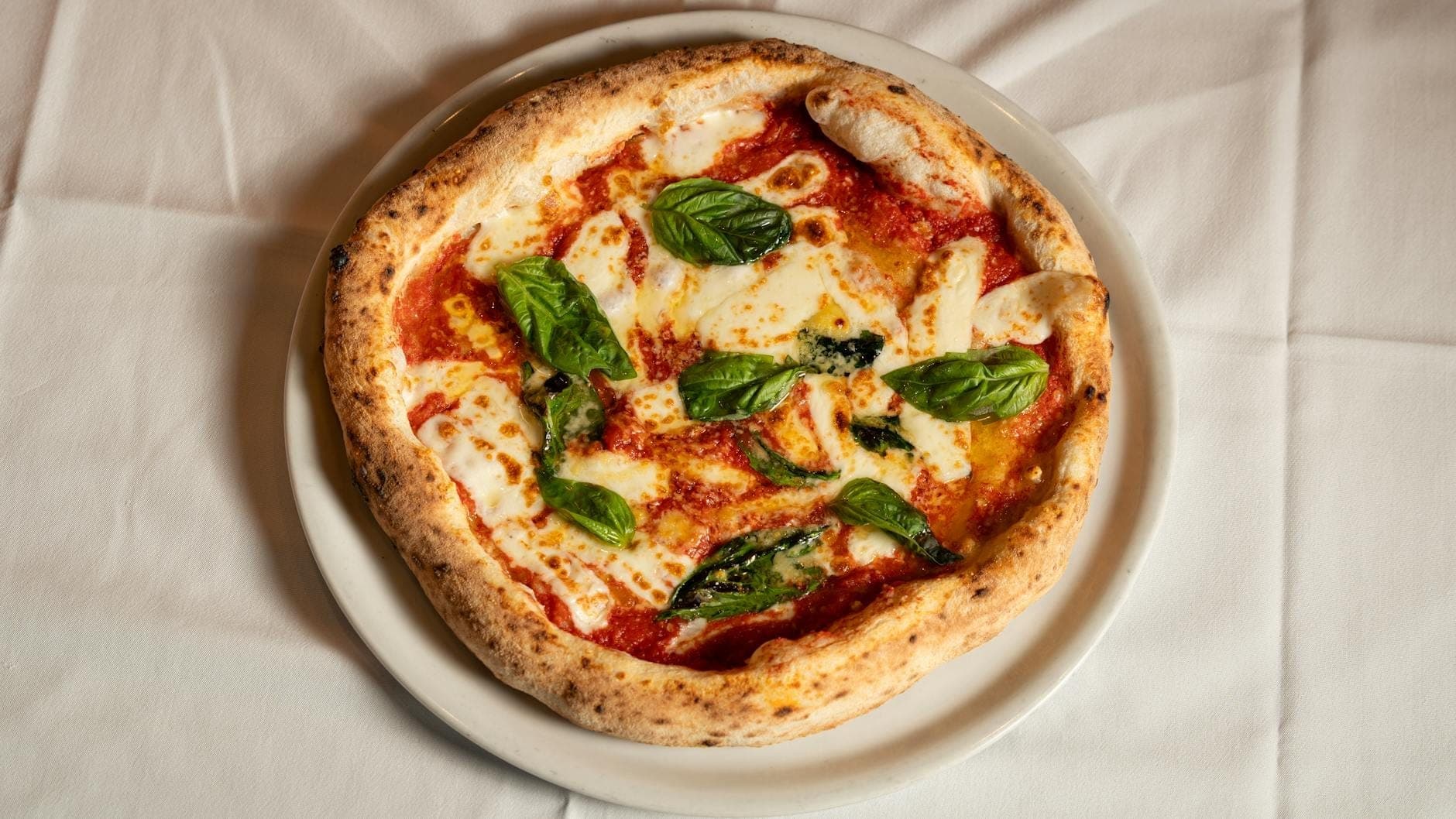 Bánh pizza margherita kiểu Napoli cổ điển với húng quế tươi, phô mai mozzarella và sốt cà chua, được bày trên đĩa trắng đặt trên khăn trải bàn trắng, nhìn từ trên xuống.