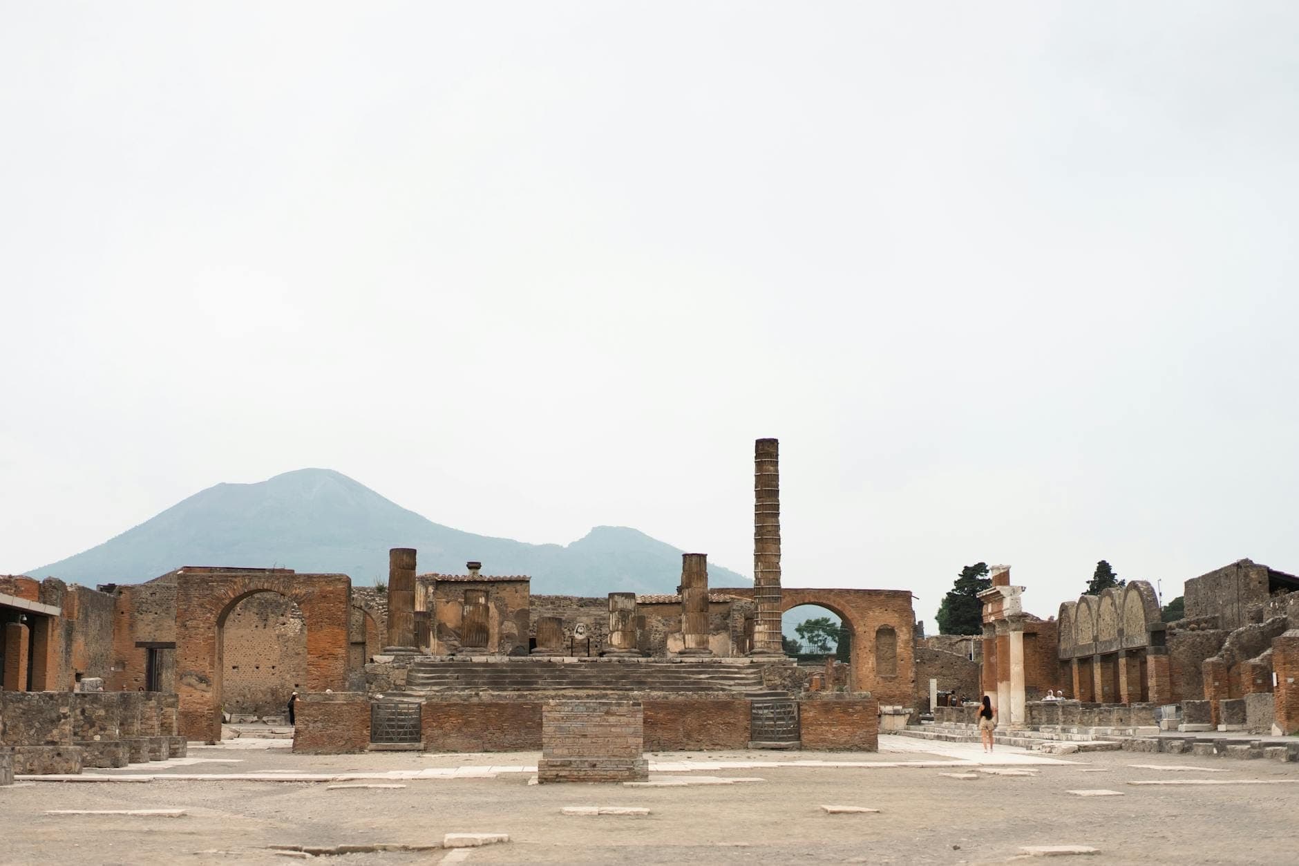 Toàn cảnh phế tích cổ đại Pompeii với núi Vesuvius ở phía sau dưới bầu trời nhạt màu, gợi lên hành trình lịch sử từ Naples đến Pompeii.