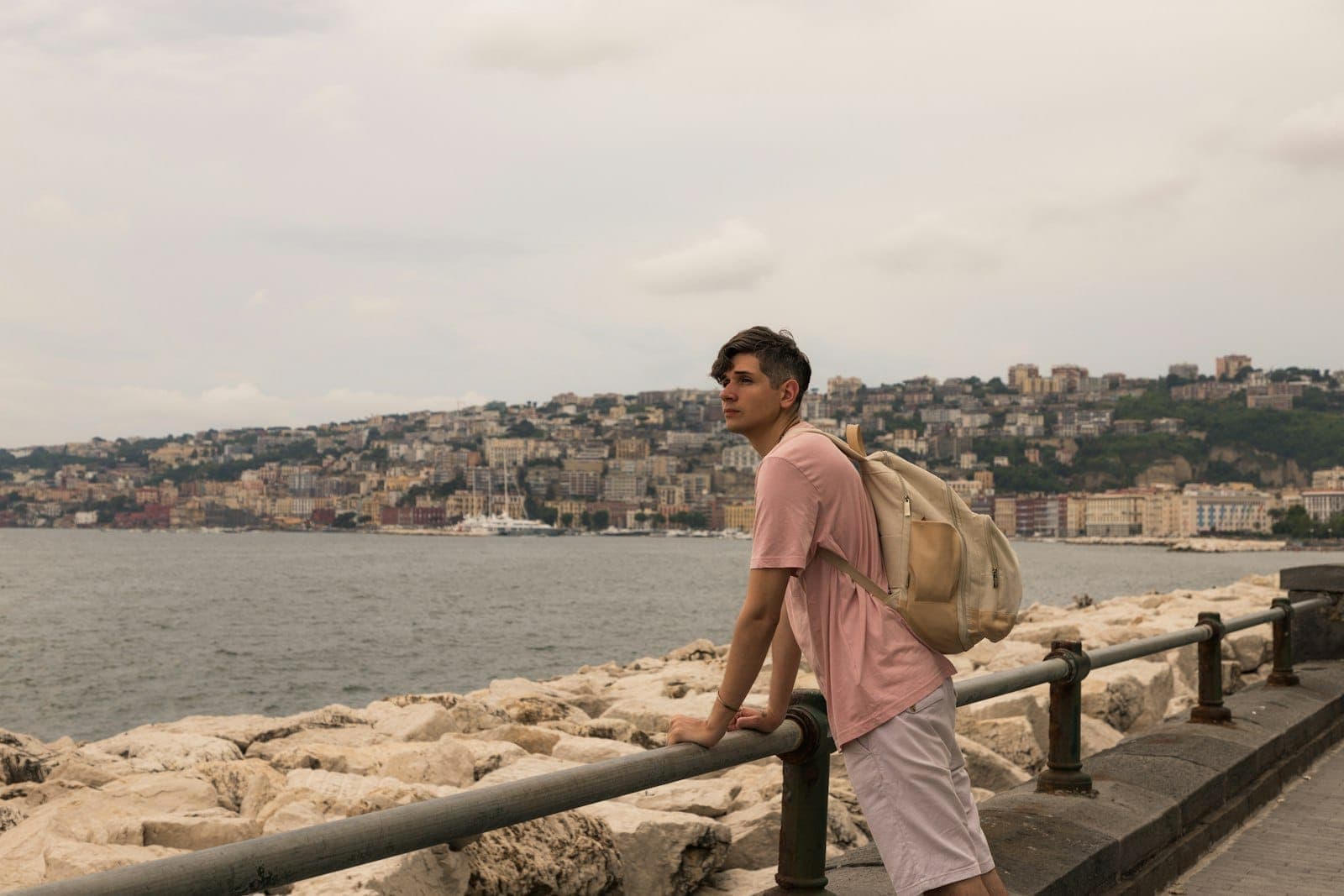 Một chàng trai trẻ đeo ba lô tựa vào lan can bờ sông ở Naples, với các tòa nhà thành phố và những ngọn đồi tối màu ở phía sau.