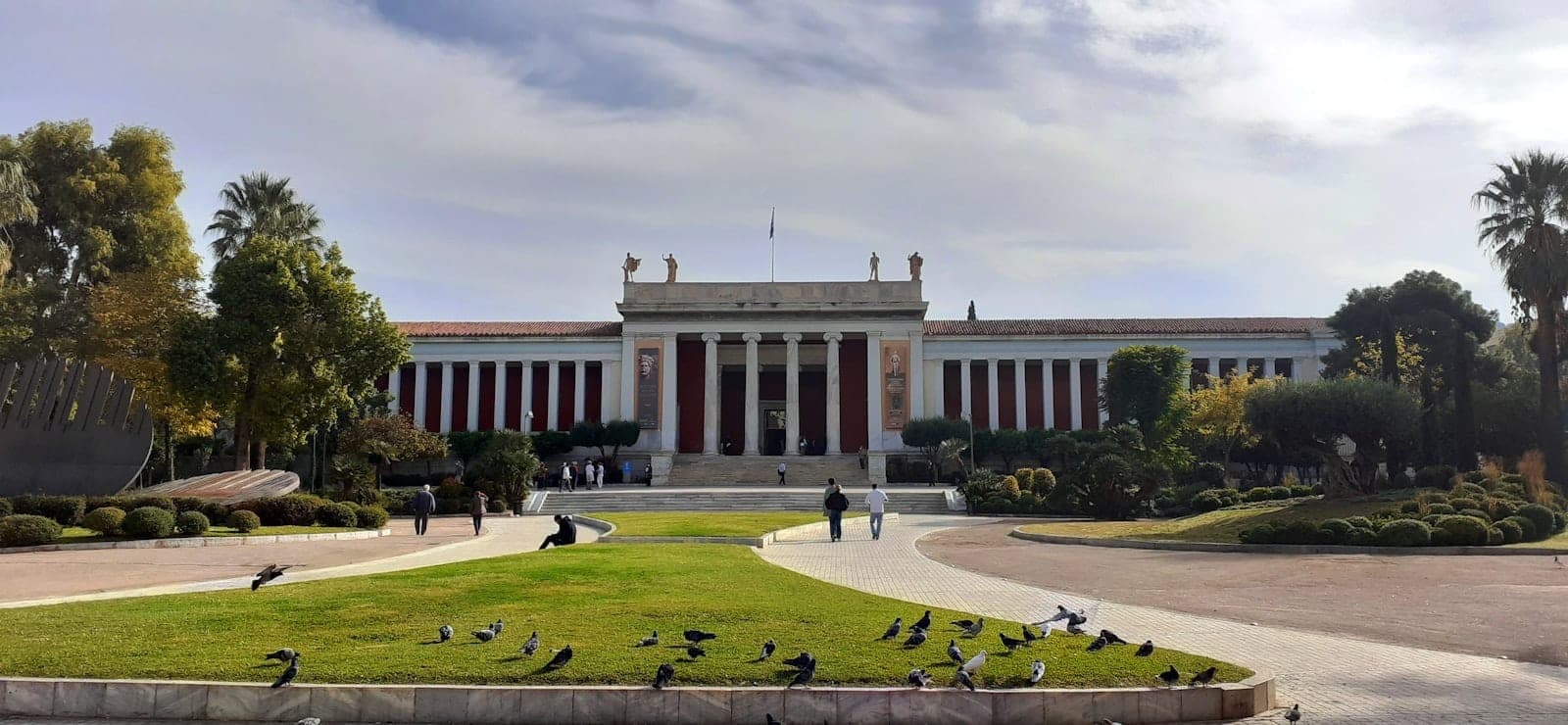 Front view of the National Archaeological Museum Athens with neoclassical columns, statues on the roof, green gardens, and people walking nearby.