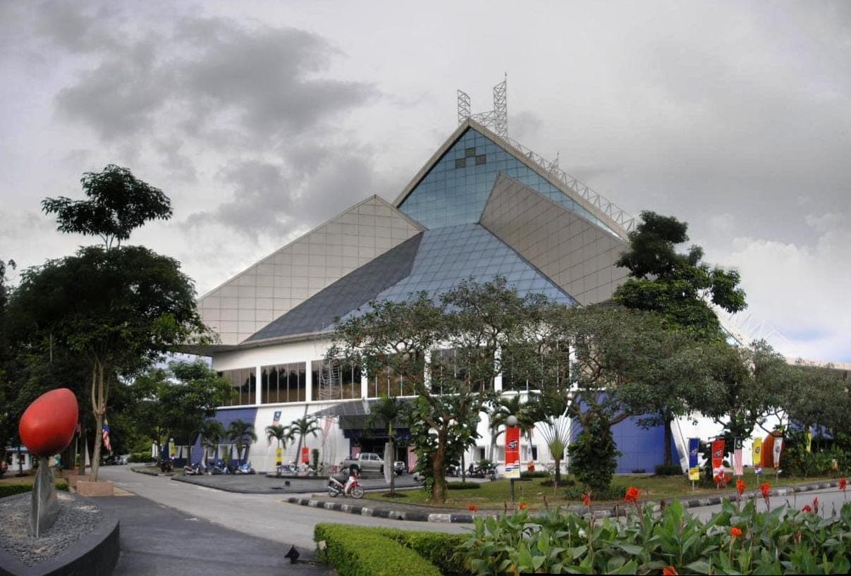 Toàn cảnh bên ngoài National Art Gallery Kuala Lumpur, nổi bật với kiến trúc hình học bằng kính và thép, cây xanh tươi tốt và bức tượng đầy màu sắc dưới bầu trời nhiều mây.