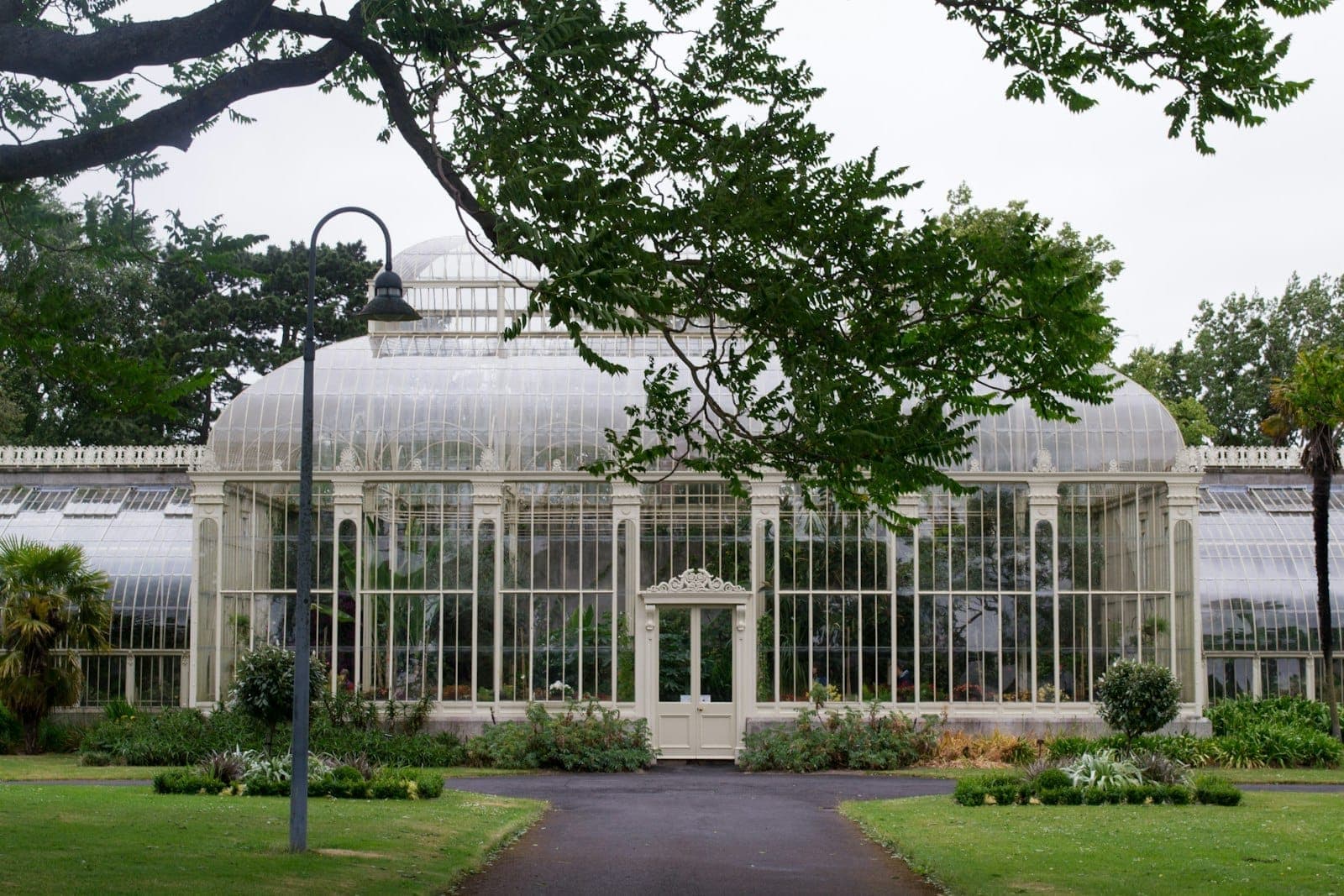 Invernadero victoriano restaurado en el Jardín Botánico Nacional de Glasnevin, rodeado de exuberante vegetación y jardines, visto desde el camino principal en un día nublado.