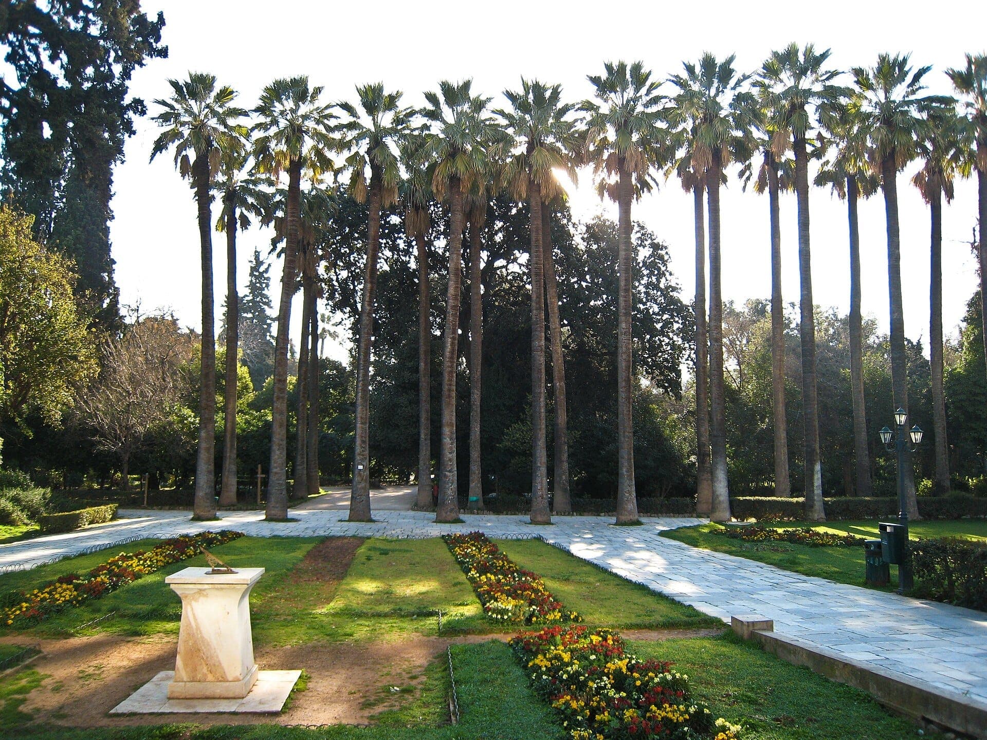 Tall palm trees, flower beds, and a stone sundial in the central walkway of the National Garden of Athens under bright sunlight.