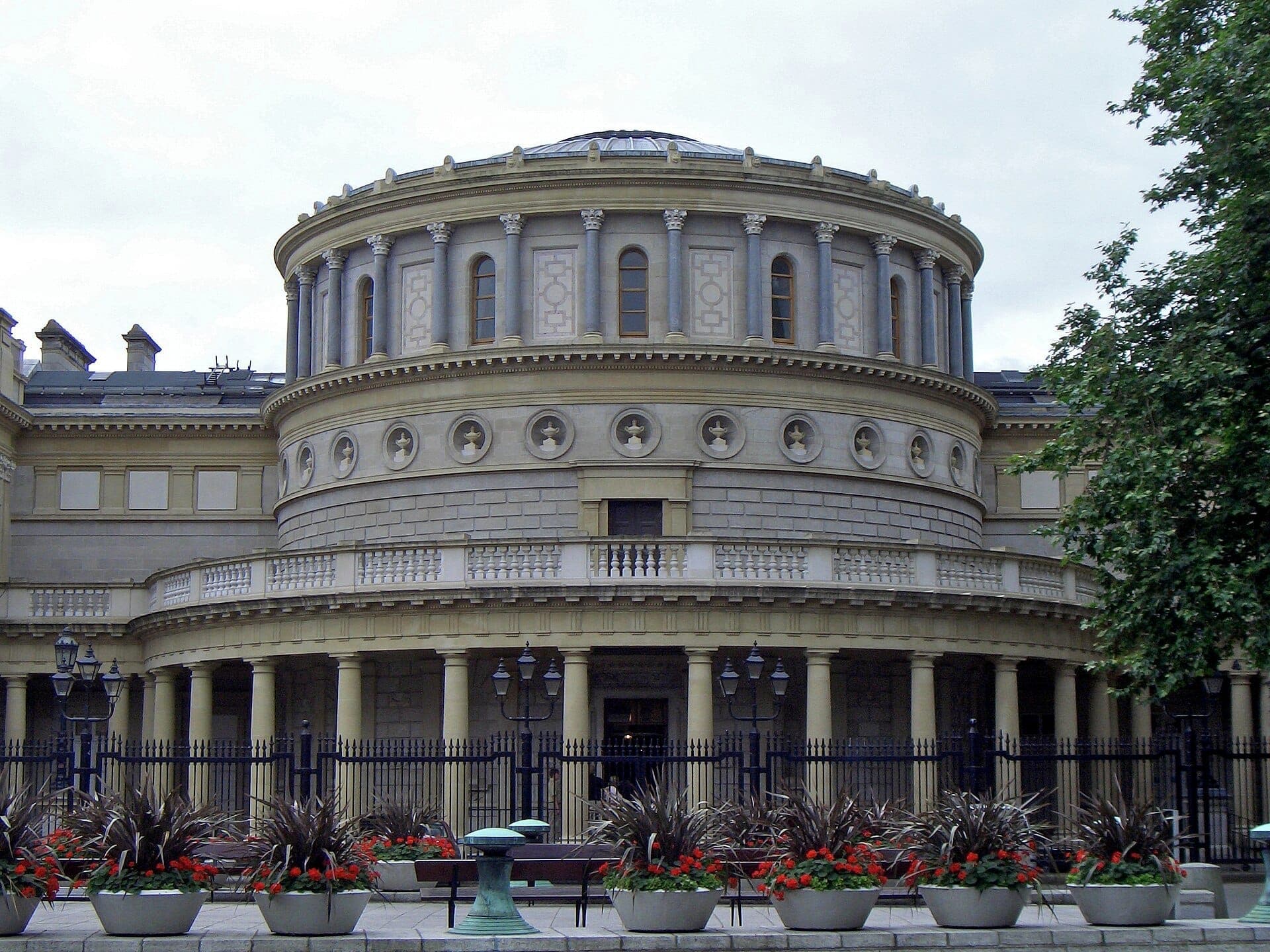 La gran rotonda y fachada neoclásica del National Museum of Ireland – Archaeology, con columnas decorativas, vallas de hierro y macetas con flores en el frente.