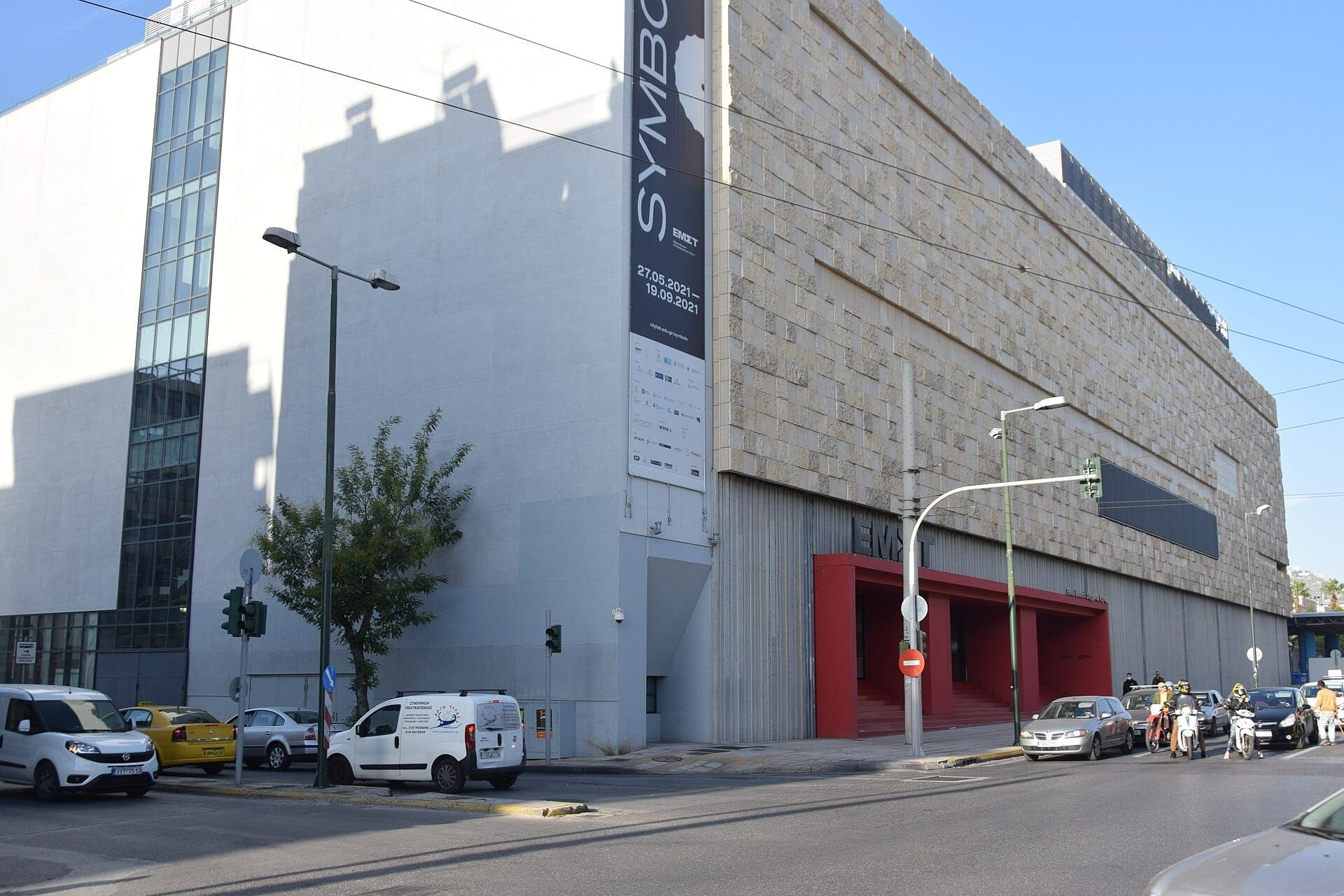 Street view of the EMST Athens building with stone and concrete exterior, cars parked along the road, and a large vertical museum banner.