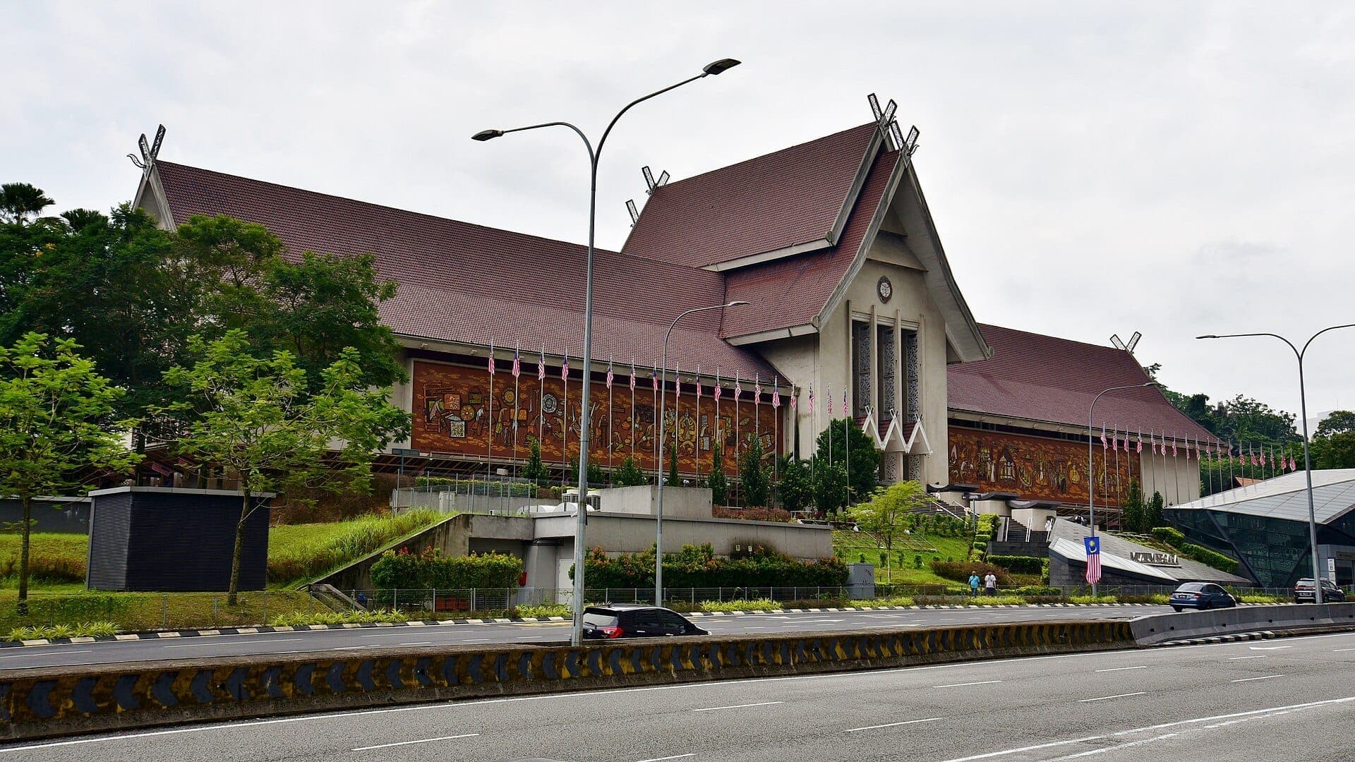 Bên ngoài National Museum Malaysia với mái dốc truyền thống, tranh tường rực rỡ và cờ Malaysia treo dọc tòa nhà vào một ngày nhiều mây tại Kuala Lumpur.