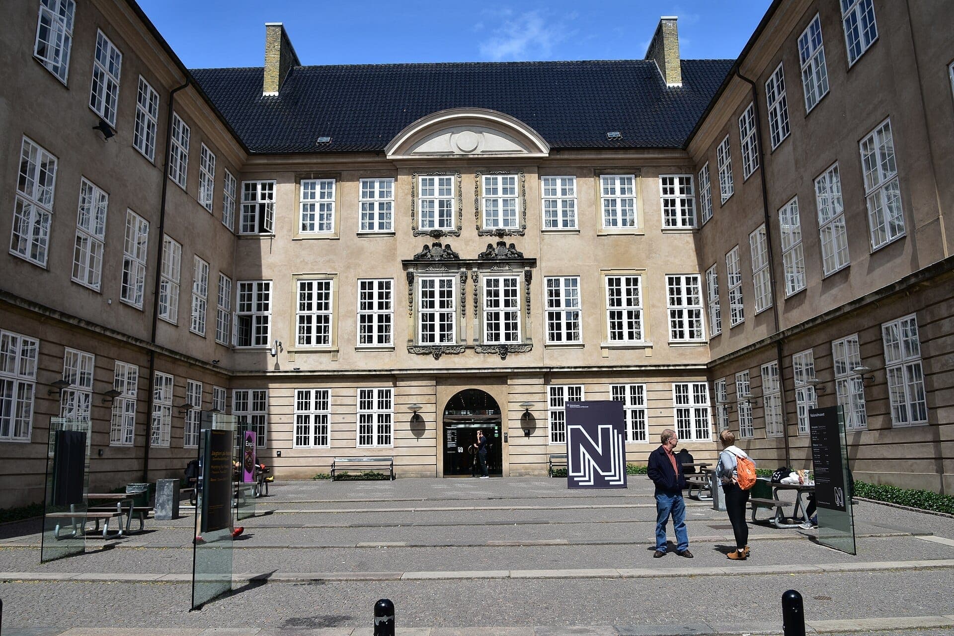 Innenhof des Nationalmuseums Dänemark mit historischer Fassade, großen Fenstern und Besuchern neben modernen Informationsschildern an einem sonnigen Tag.